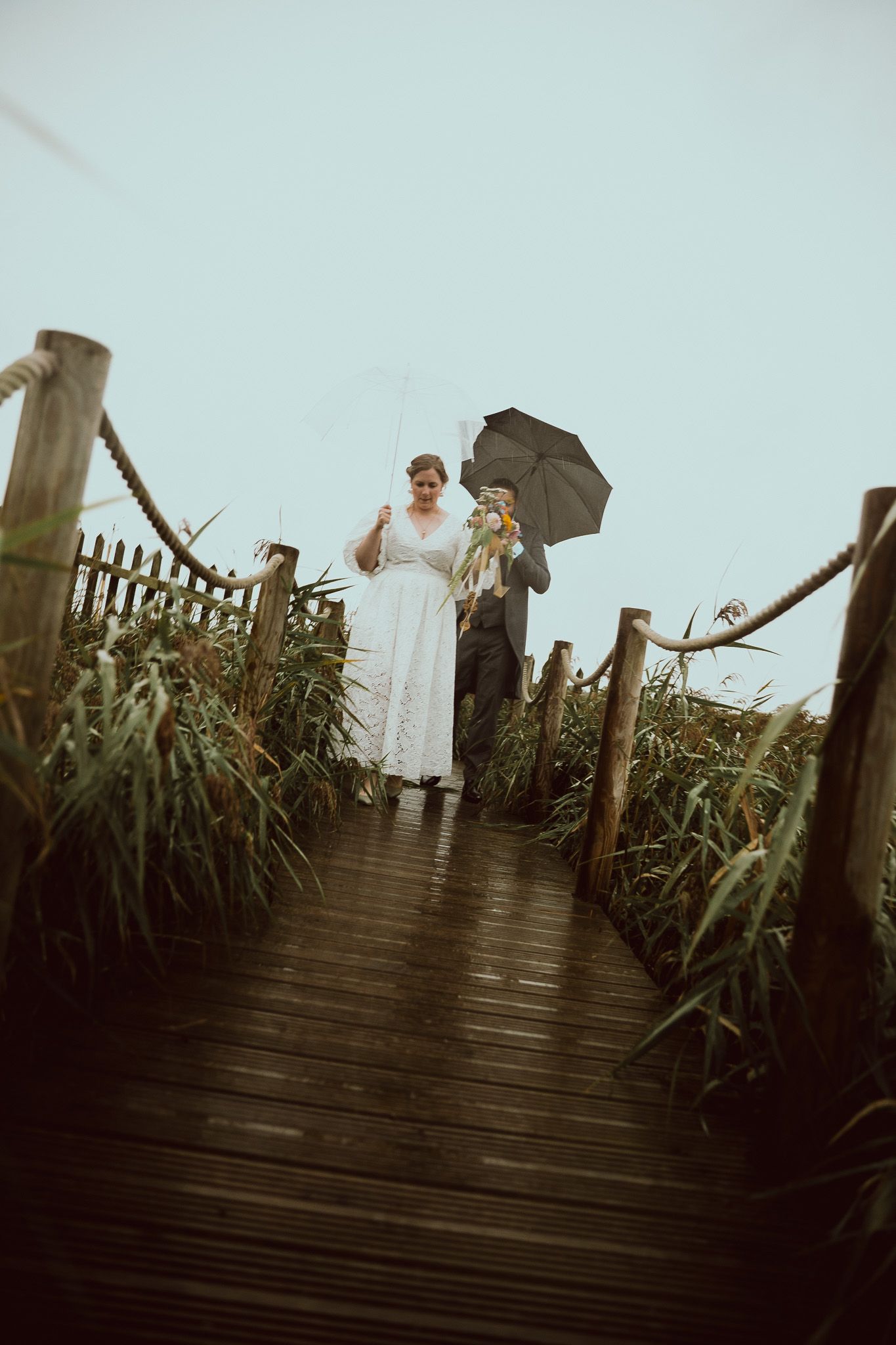 People walking on a wooden boardwalk through tall grass on an overcast day with black umbrellas.