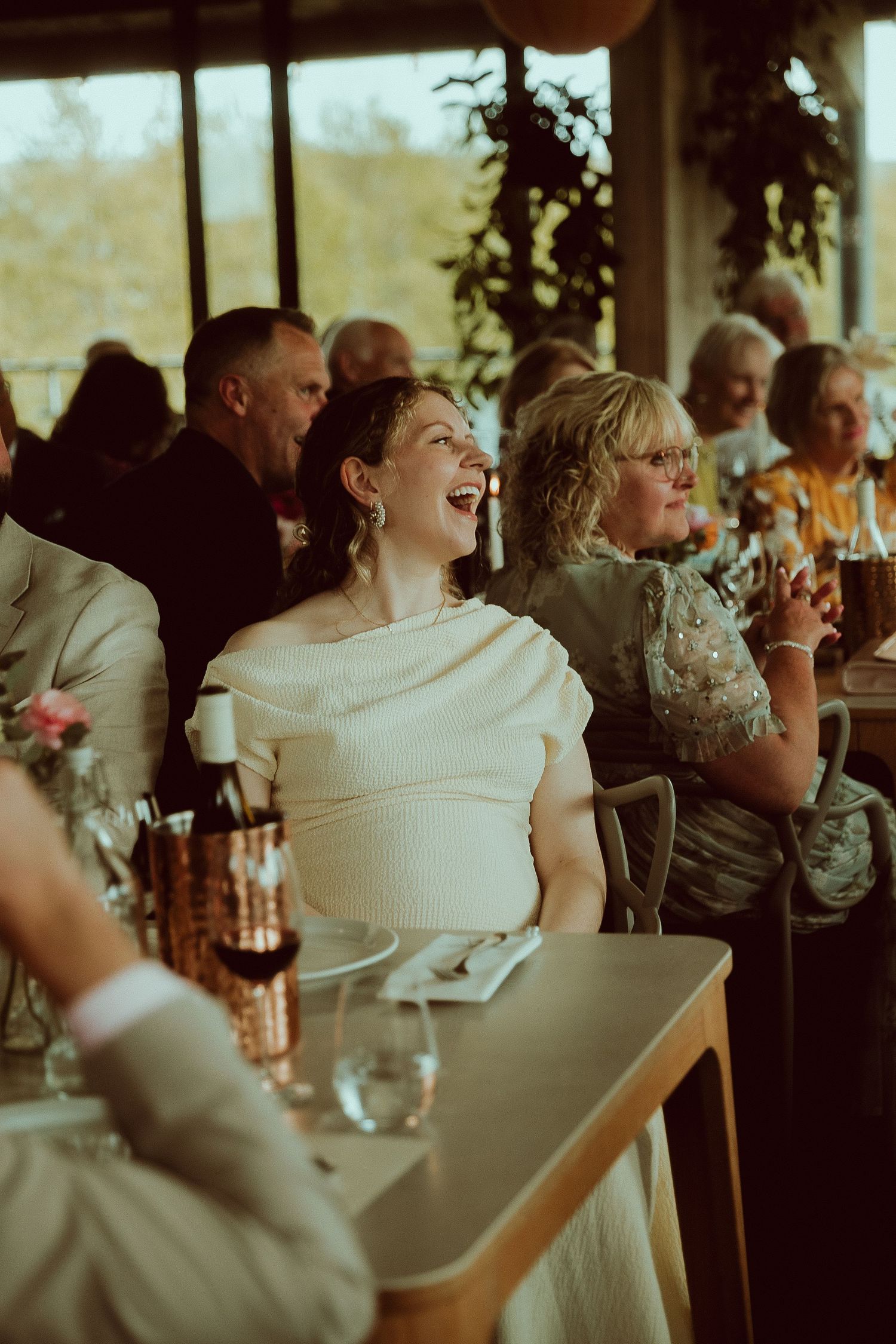 A laughing wedding guest in white sits at a wedding reception table surrounded by smiling guests in a bright venue.
