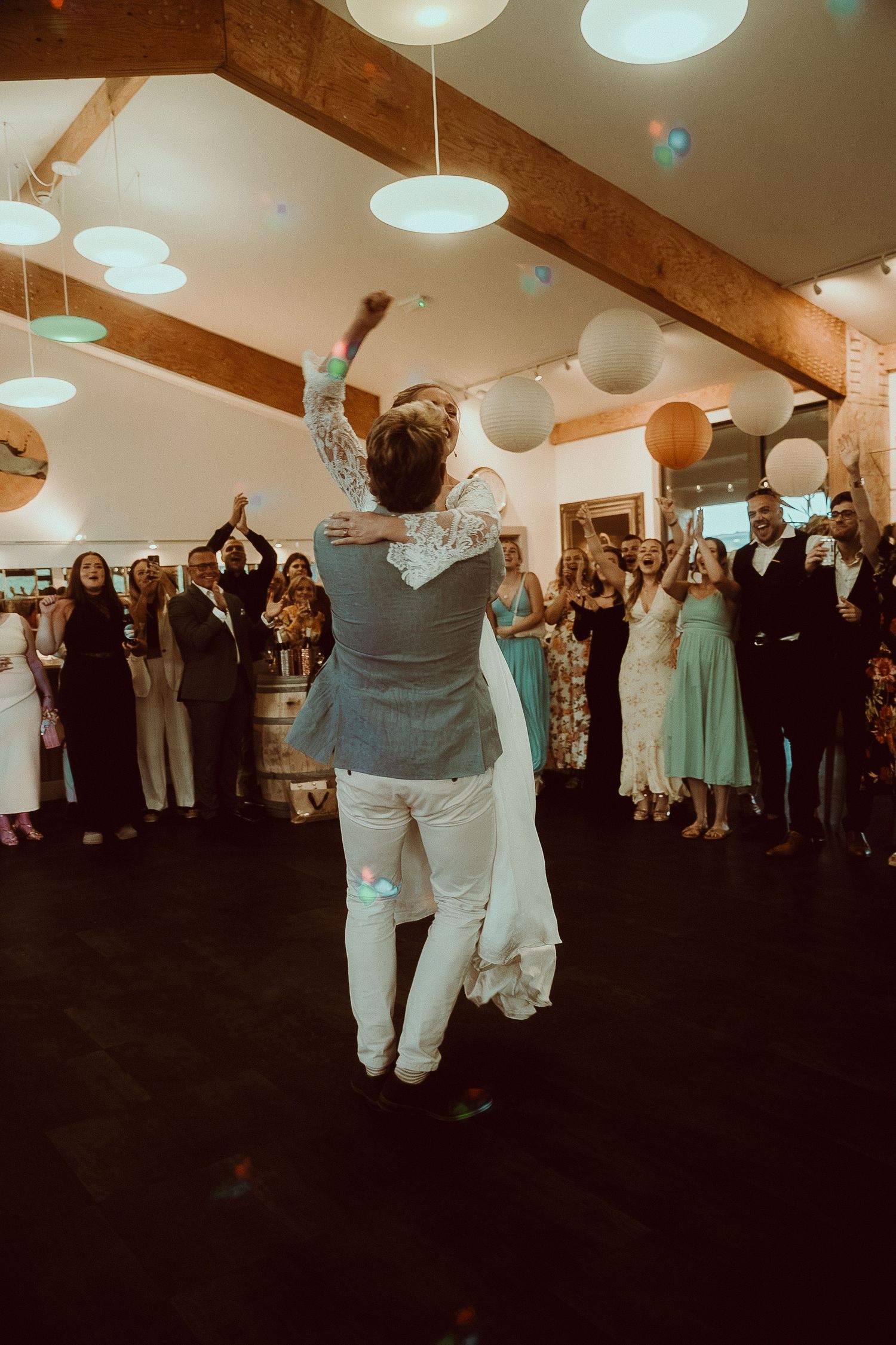 Couple sharing their first dance at a wedding reception while guests cheer and applaud in a warmly lit venue.