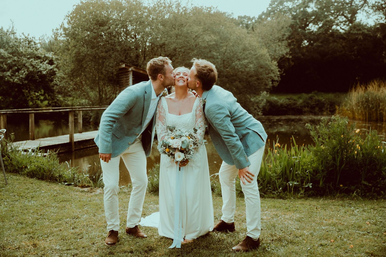 Bride in white gown kissed on cheeks by two groomsmen in blue jackets outdoors near a pond.