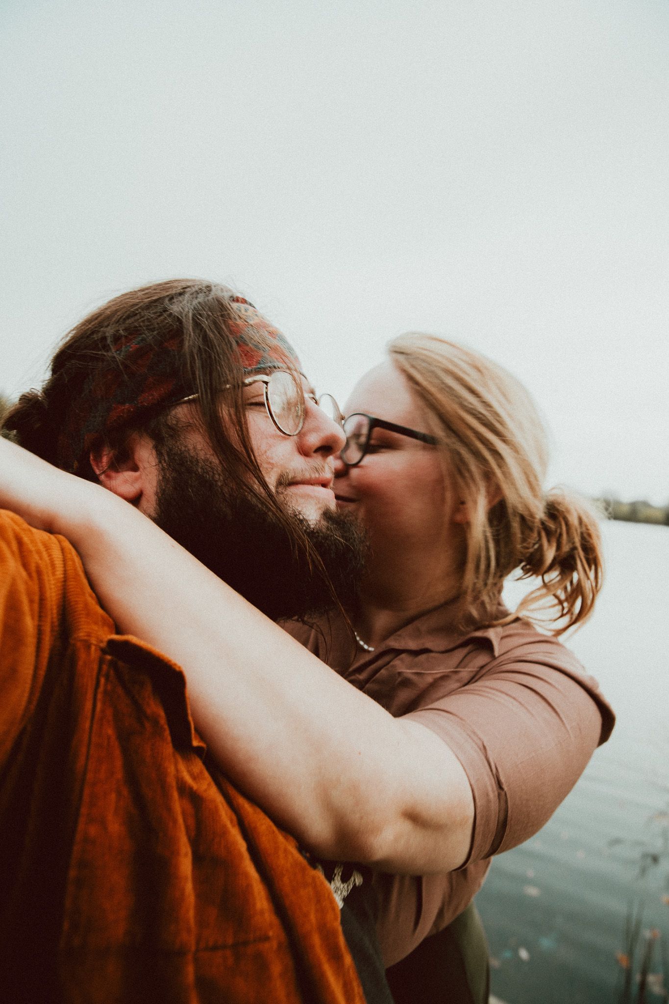 A bearded man and blonde woman with glasses share a romantic kiss outdoors near water.