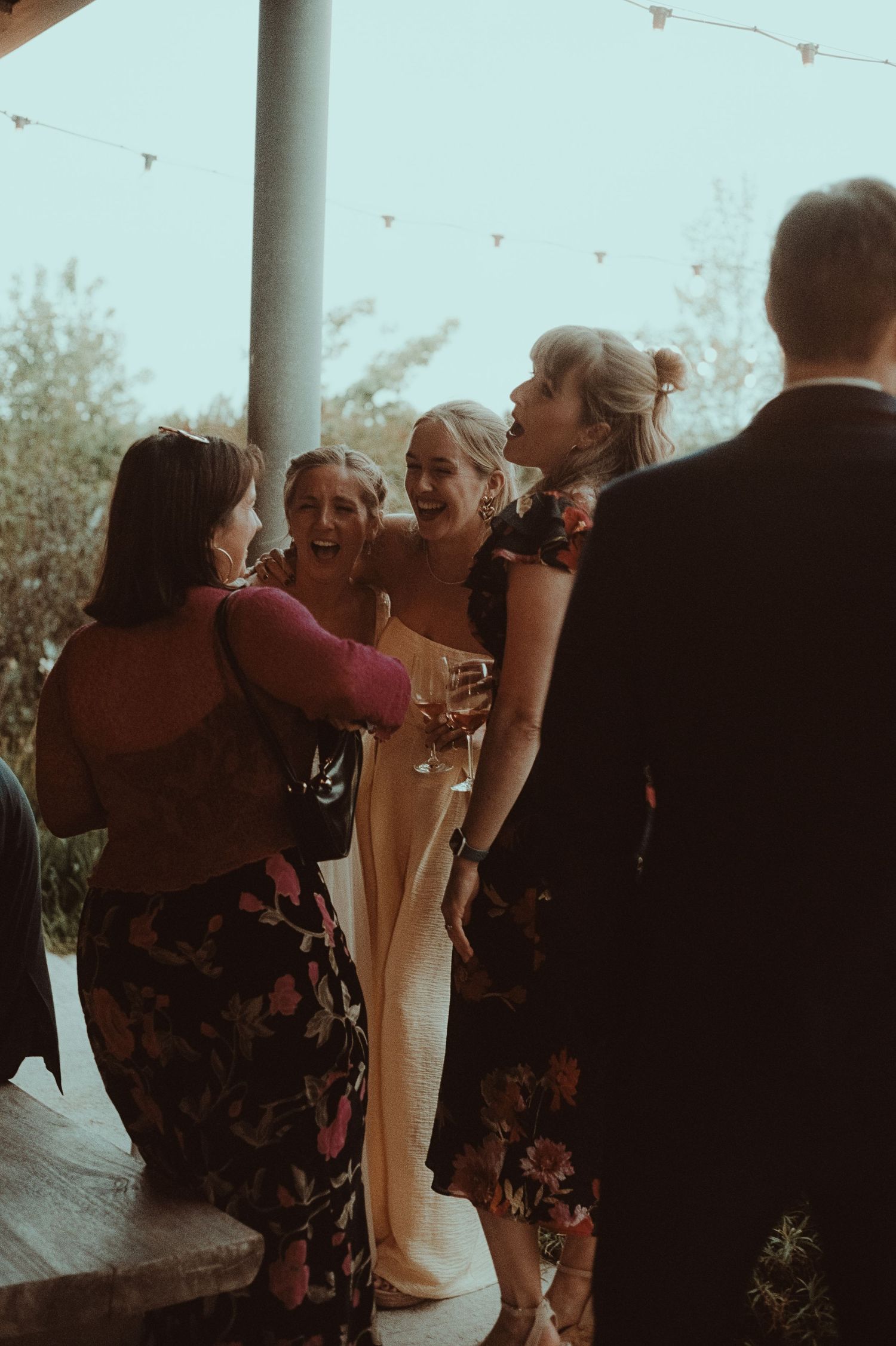 Bride laughing with bridesmaids in floral dresses at an outdoor wedding reception, holding champagne glasses.