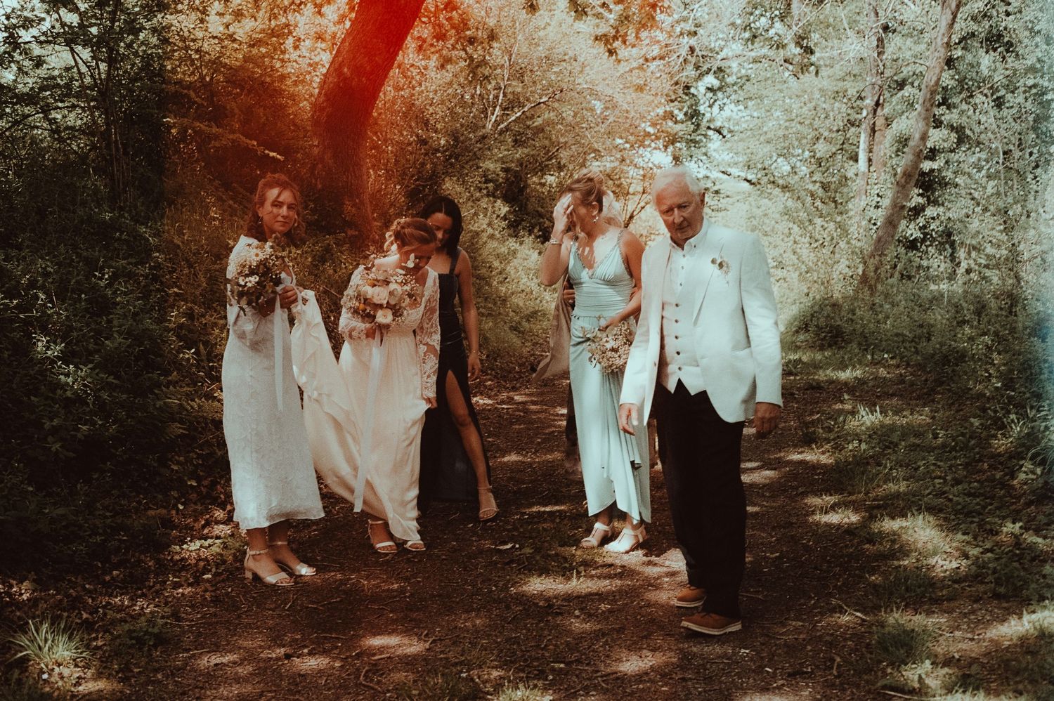 Wedding party walking on a forest path, brides in white gowns with bouquets, groom in white suit.