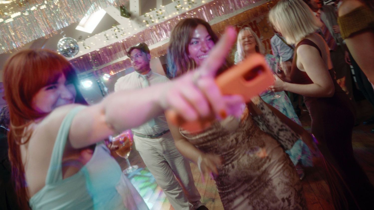 People dancing at a lively party with disco ball lights, a woman pointing a pink toy gun at the camera.