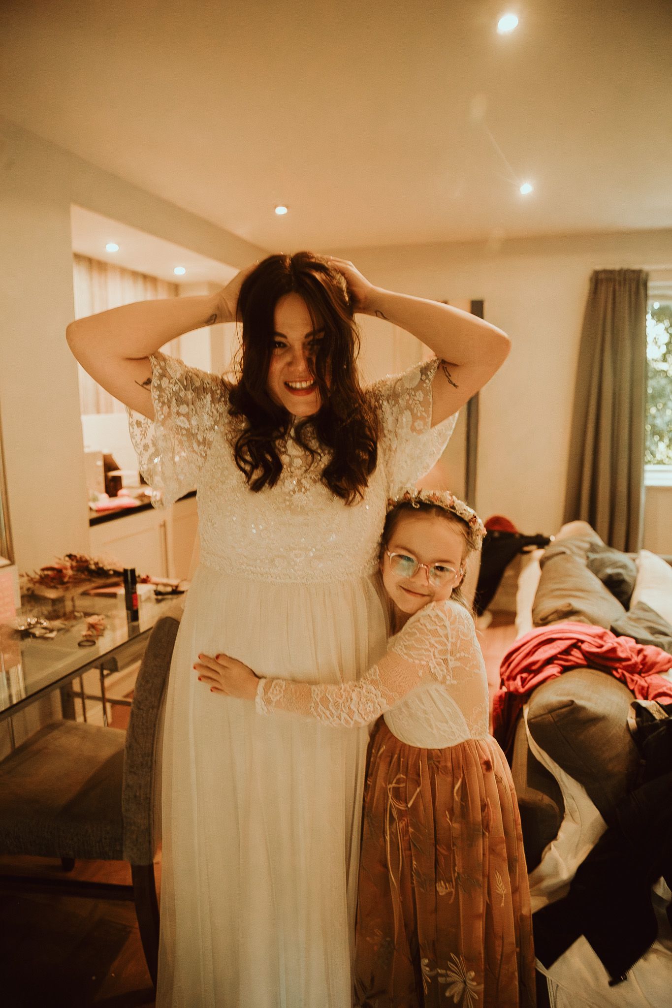 Bride in white lace wedding dress smiling while young flower girl in tulle skirt hugs her during bridal preparations.