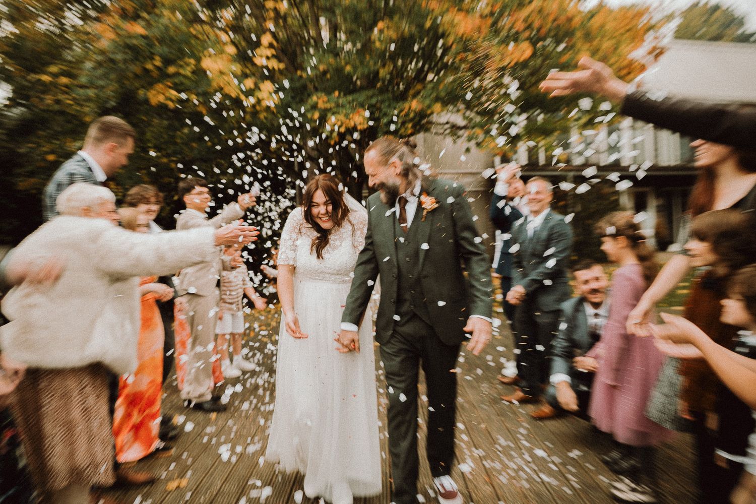 Bride and groom walk through confetti shower surrounded by cheering wedding guests outdoors in autumn setting.