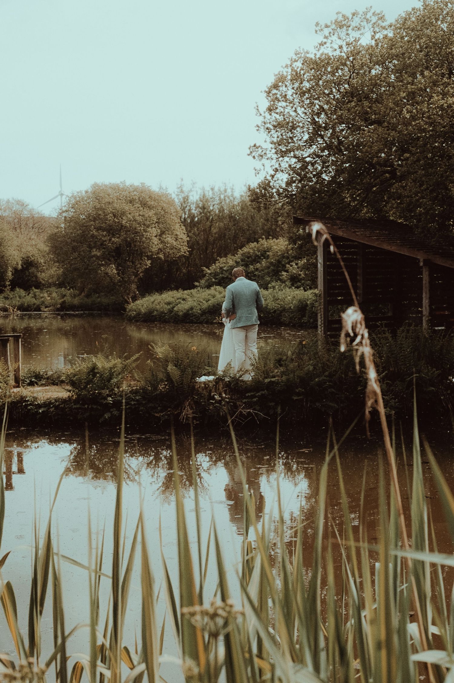 Person in formal attire standing by a tranquil pond surrounded by lush green trees and reeds.