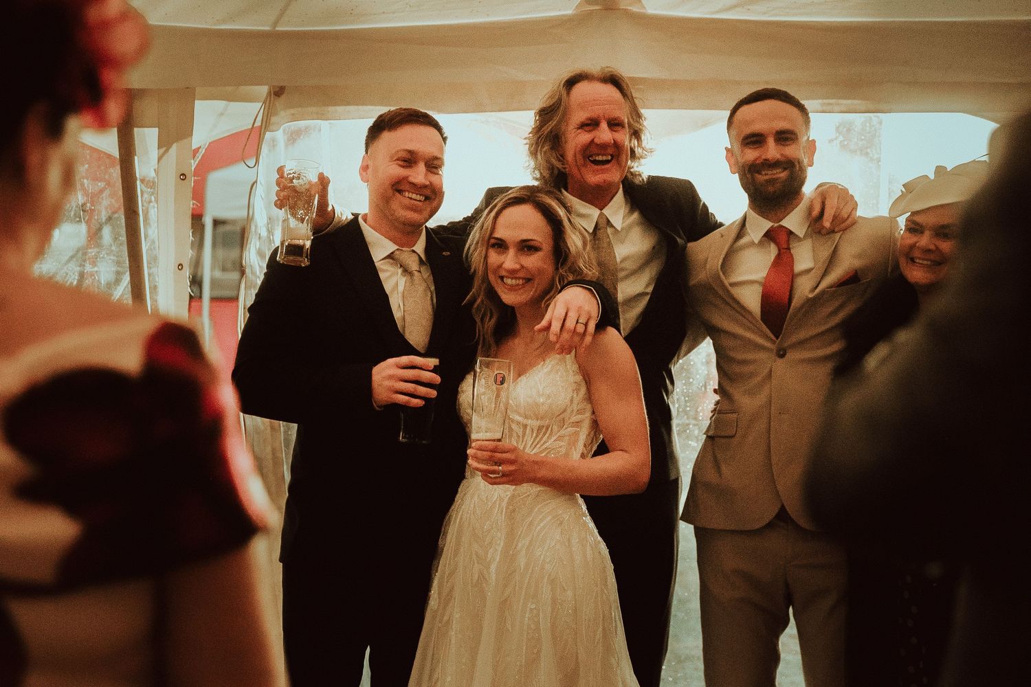 Bride and groom celebrate with guests at wedding reception, smiling and holding drinks in a marquee.