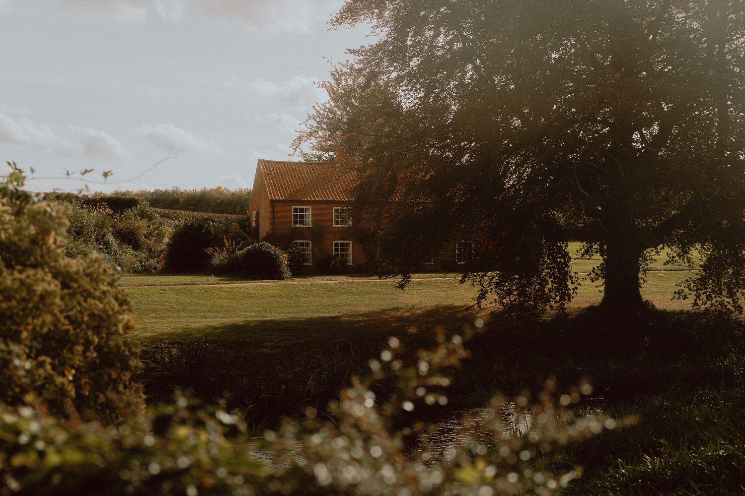 A charming red brick farmhouse surrounded by lush green lawns and mature trees in golden evening light.