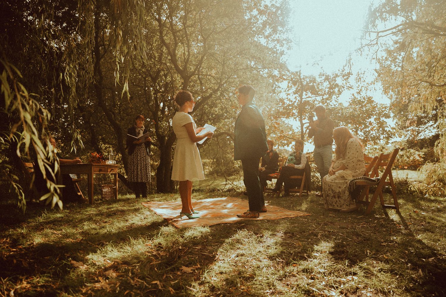 Outdoor wedding ceremony in a sunlit garden with guests gathered among trees in warm golden light.