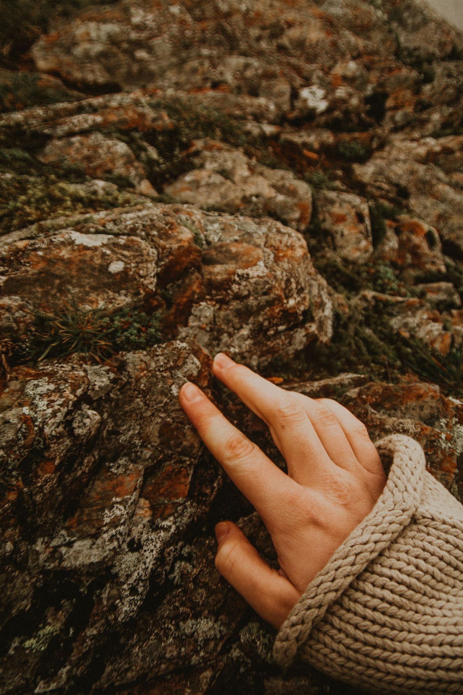 A hand in a knit sweater sleeve gently touches a rugged, lichen-covered rock surface outdoors in Scotland.