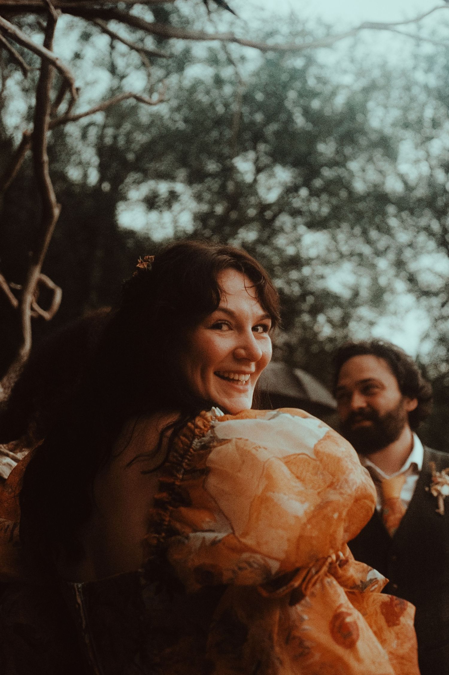 Smiling bride in orange floral dress outdoors with groom in background among trees at golden hour.
