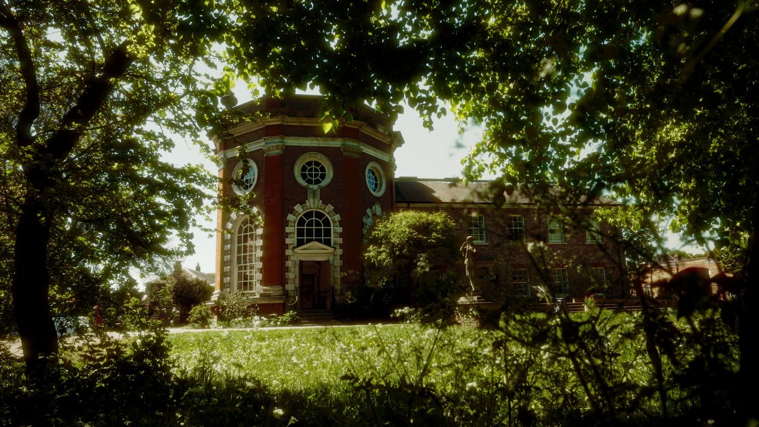 Historic red brick building with ornate architecture framed by lush green trees in a scenic garden setting.