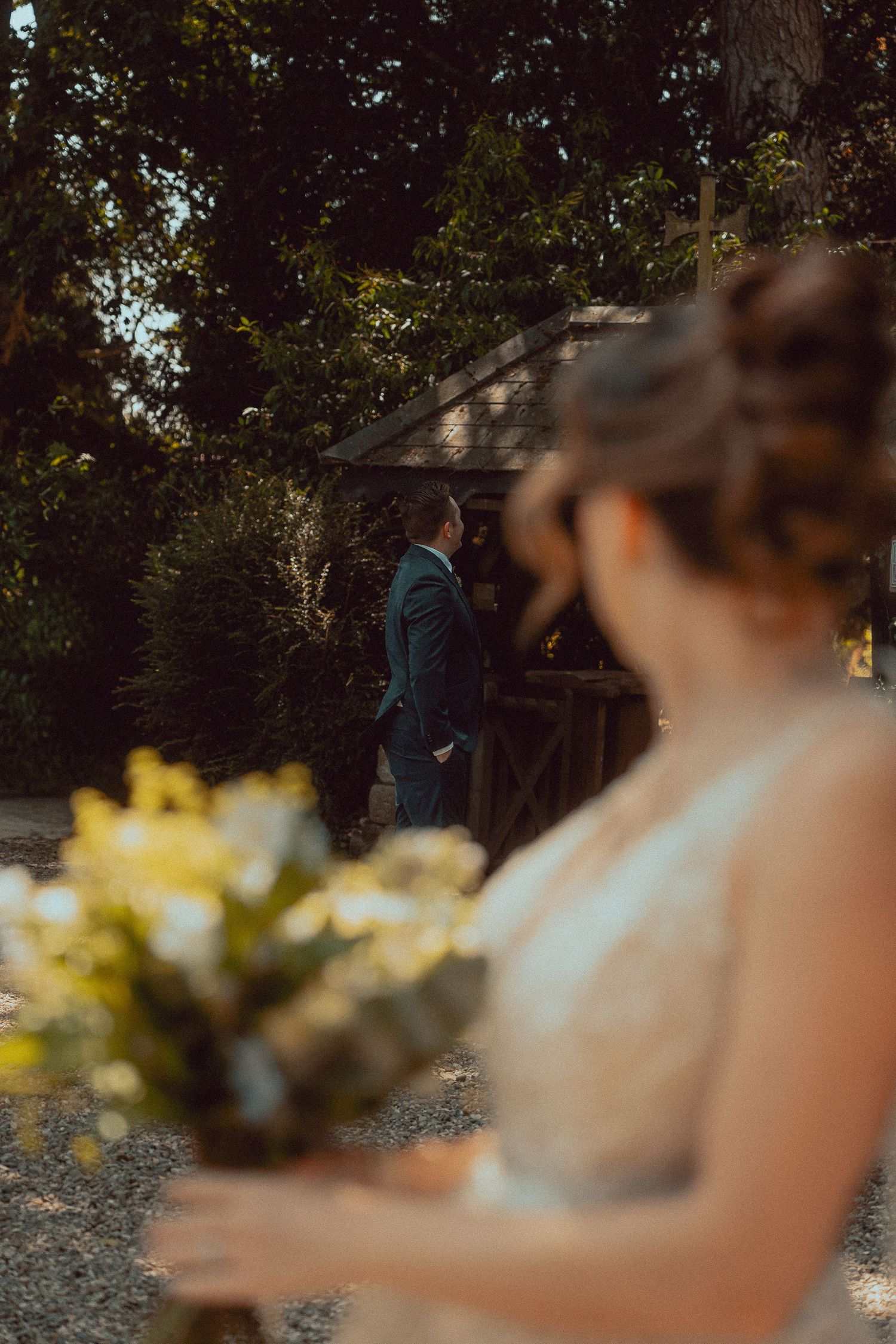 Bride holding yellow bouquet in foreground as groom walks toward her at outdoor wedding venue surrounded by trees.
