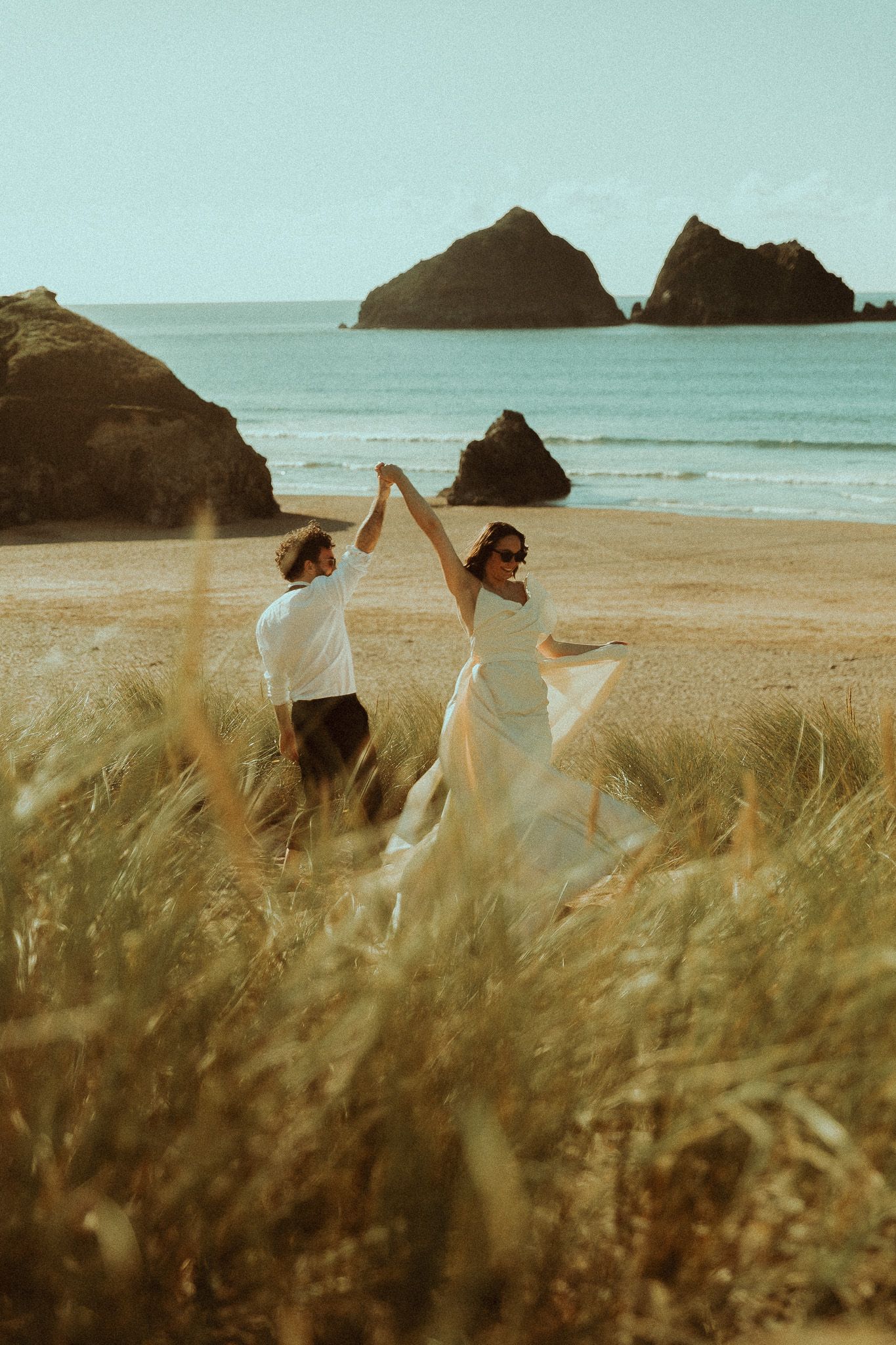 A couple dances on a sandy beach with rocky outcrops and blue ocean waves, the bride in a flowing white gown.