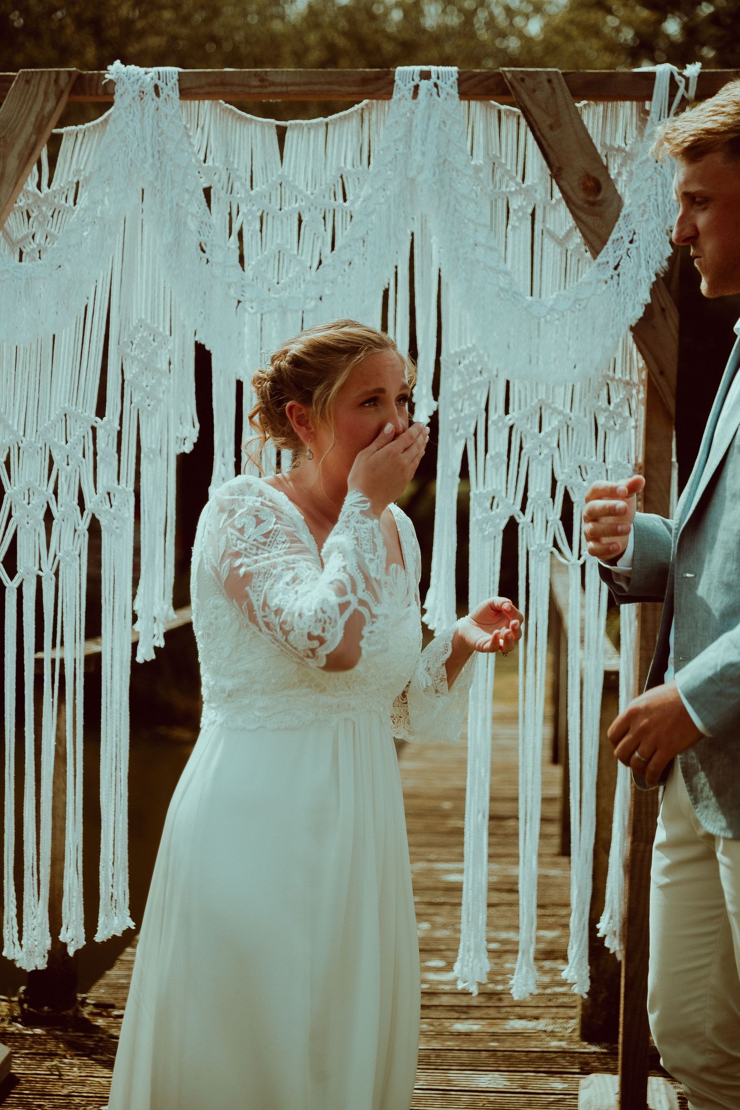 Emotional bride in lace wedding dress laughing at altar with macrame backdrop during outdoor wedding ceremony.