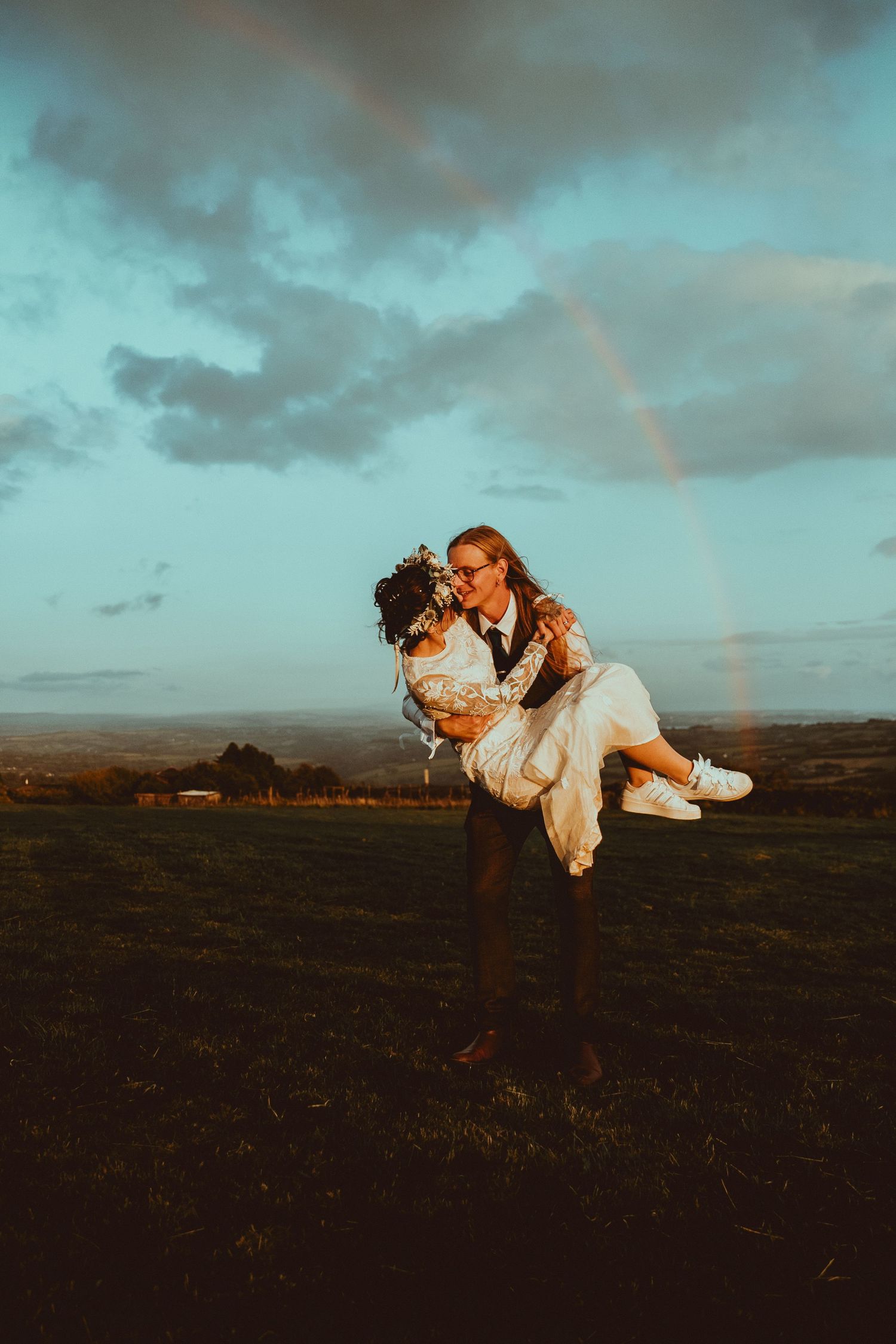 Groom lifting bride in white dress outdoors at golden hour with rainbow in cloudy sky behind them.