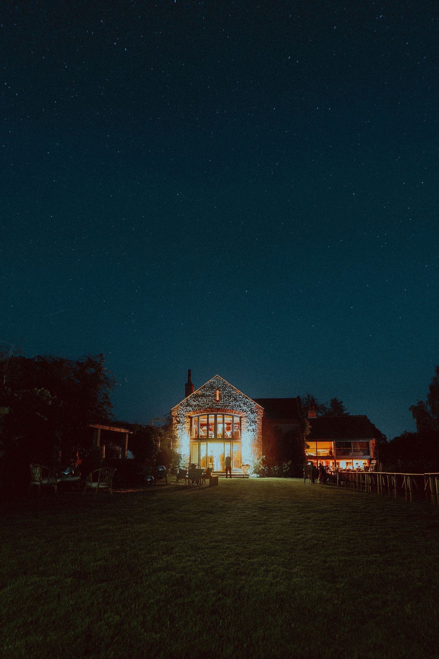 A illuminated stone barn glows warmly under a stunning starry night sky surrounded by trees and green lawn.