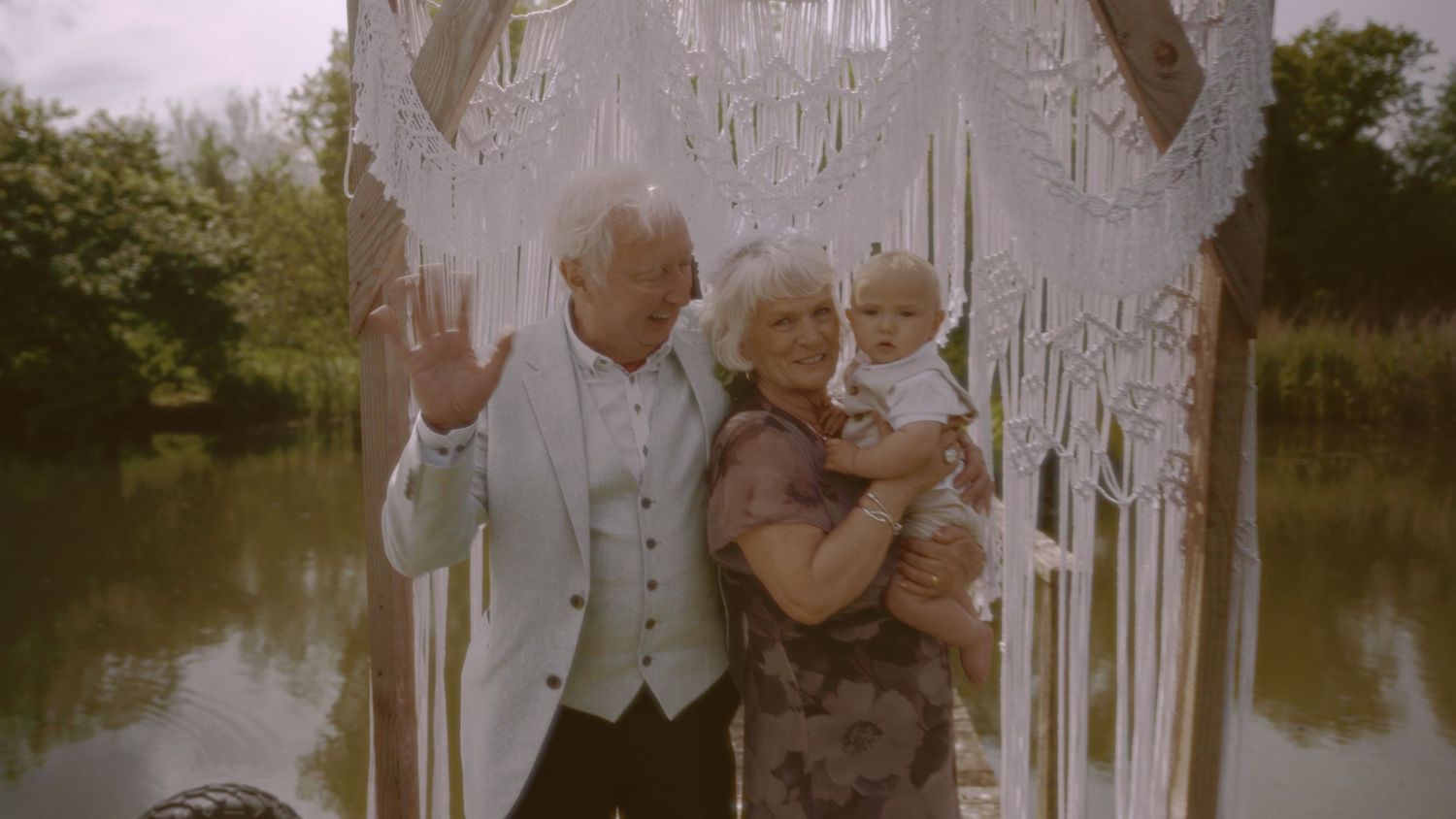 Elderly couple holding a baby near a decorative macrame arch by a lake, smiling and waving outdoors.