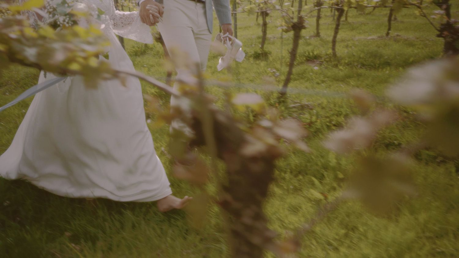 Bride and groom walking hand in hand through a lush green orchard with blooming spring trees.