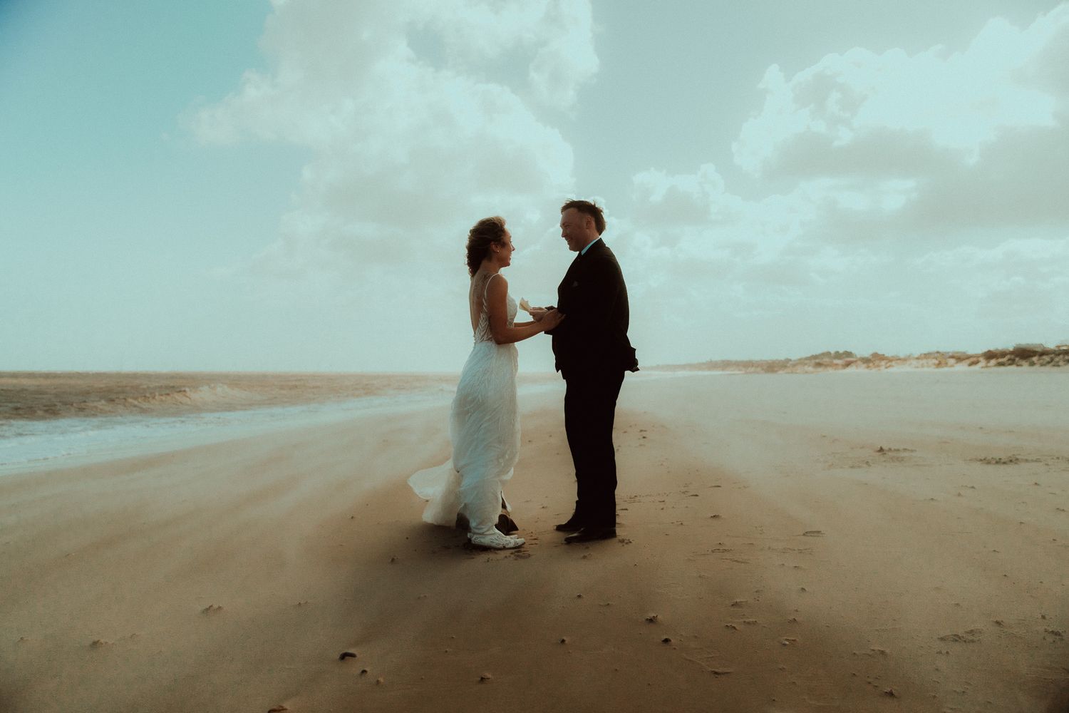 A couple exchanges vows on the Norfolk coast, bride in white gown and the groom in dark suit  during their elopement
