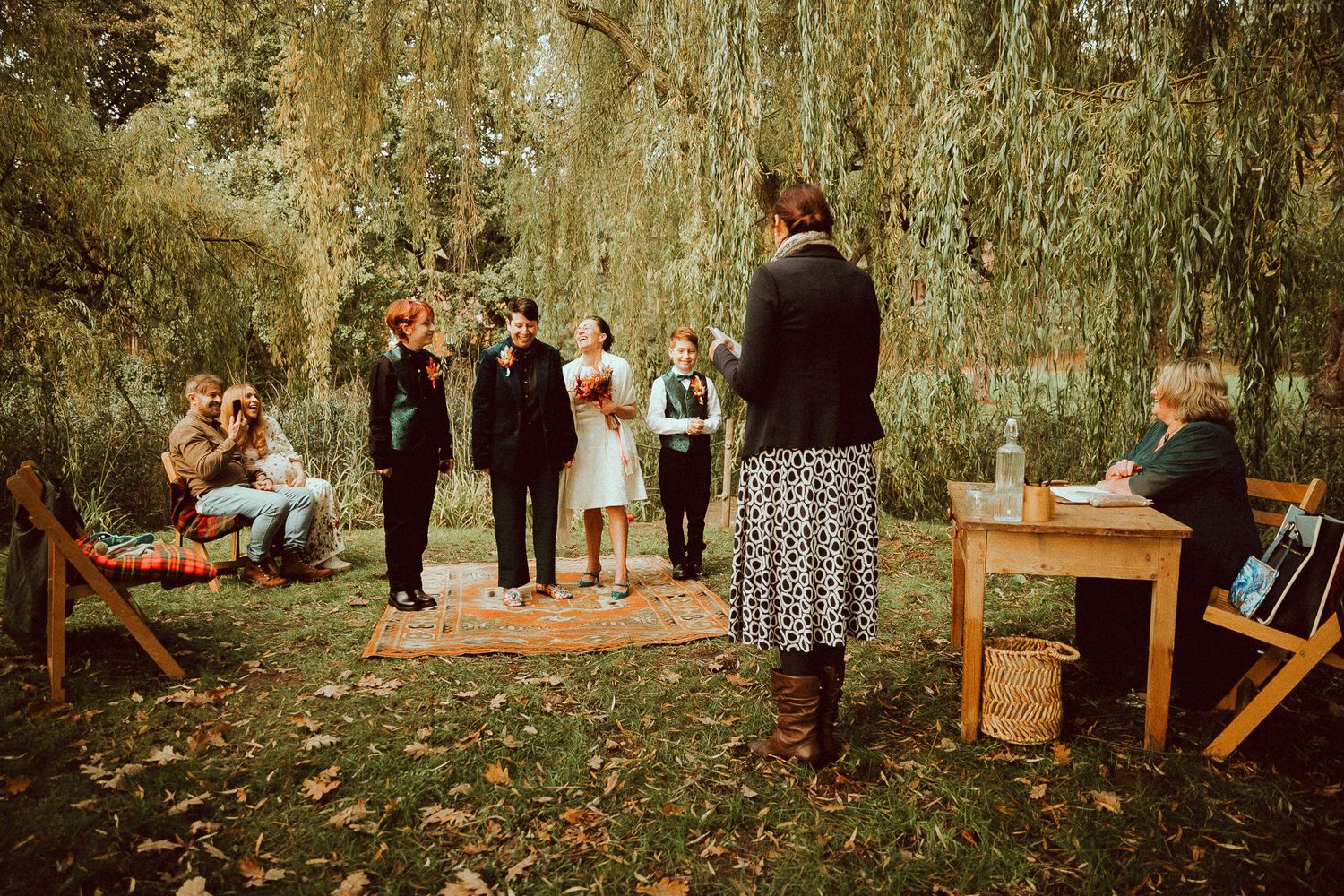 Outdoor wedding ceremony under weeping willow tree with couple, officiant, and guests on autumn day.