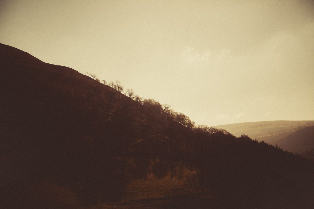 Sepia-toned landscape of rolling hills with sparse trees silhouetted against a pale sky at dusk in Wales.
