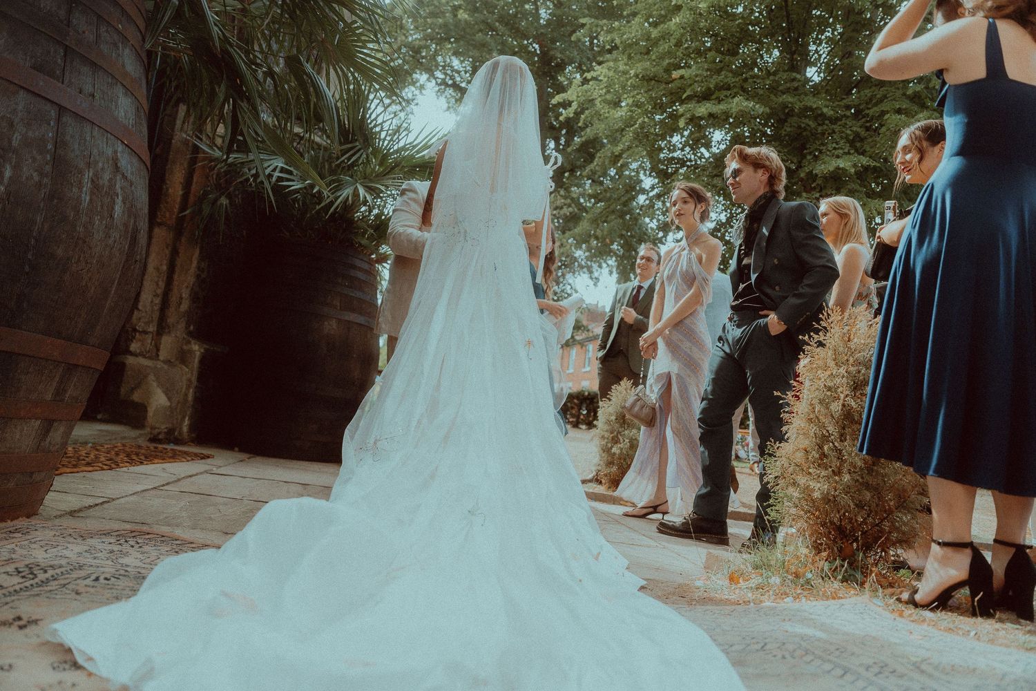 Bride in white gown and veil stands at outdoor wedding ceremony surrounded by guests and lush greenery.