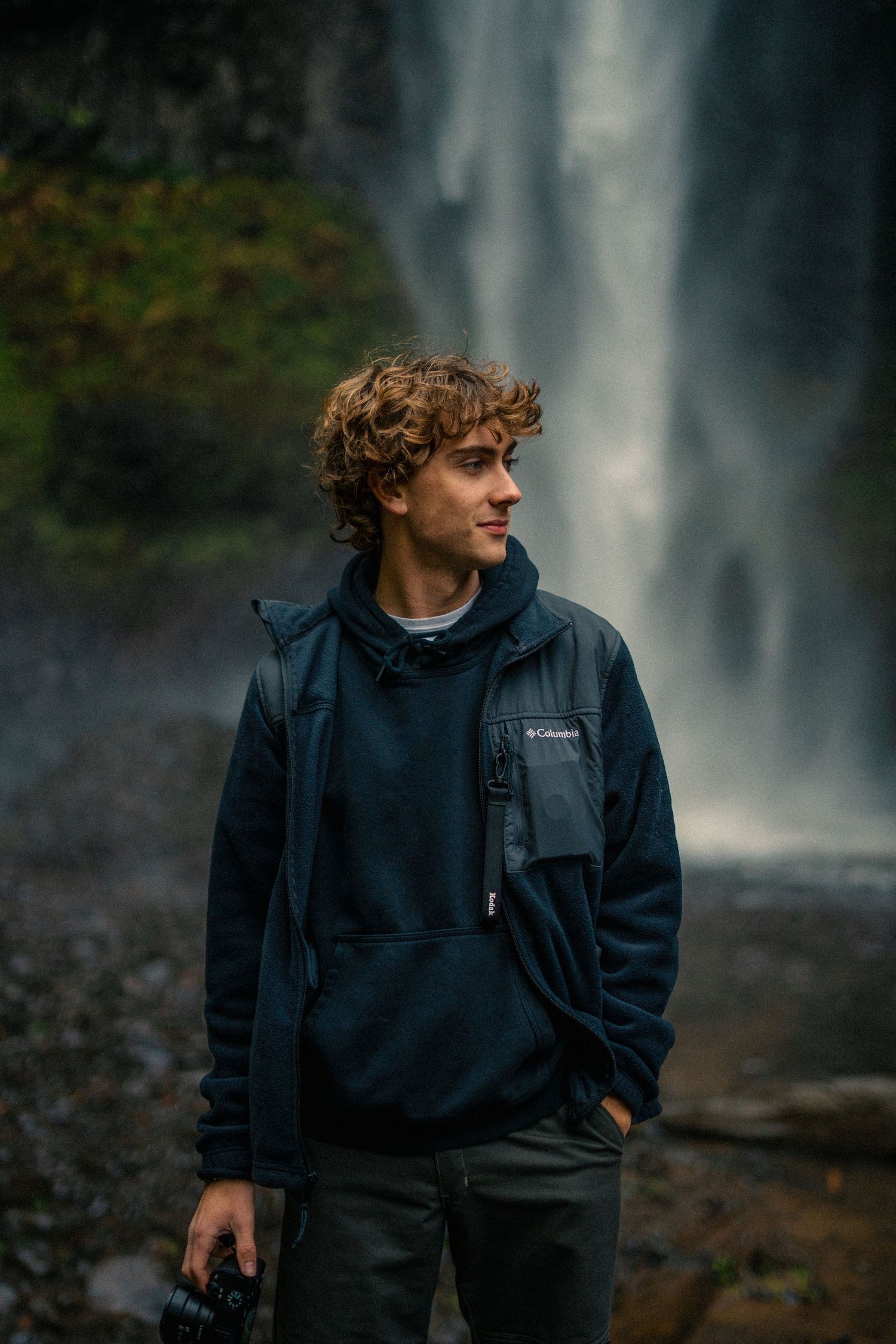 Person in navy hoodie standing near majestic waterfall in moody natural setting.