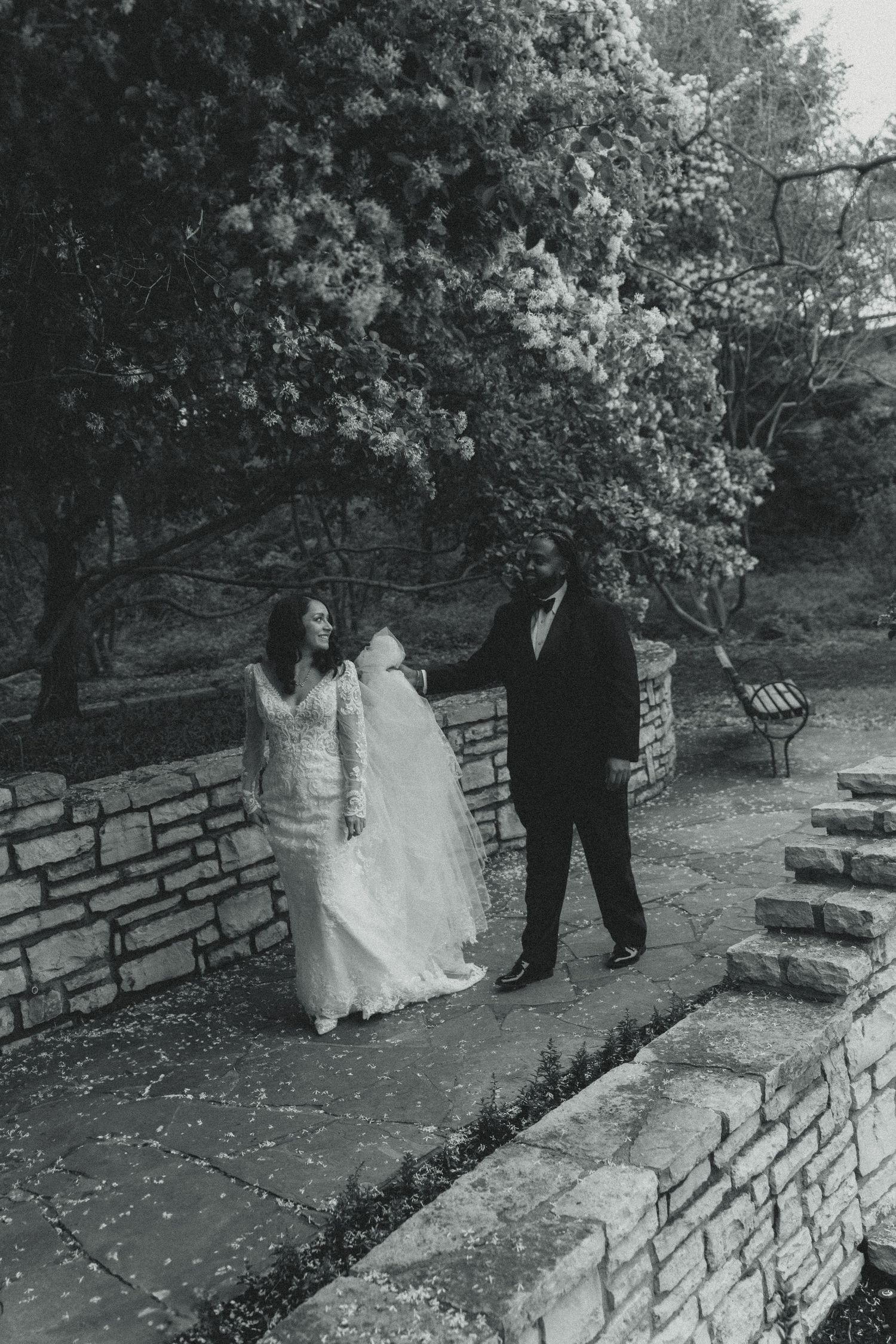 Black and white photo of a couple in wedding attire standing on stone steps near flowering trees in a garden setting.