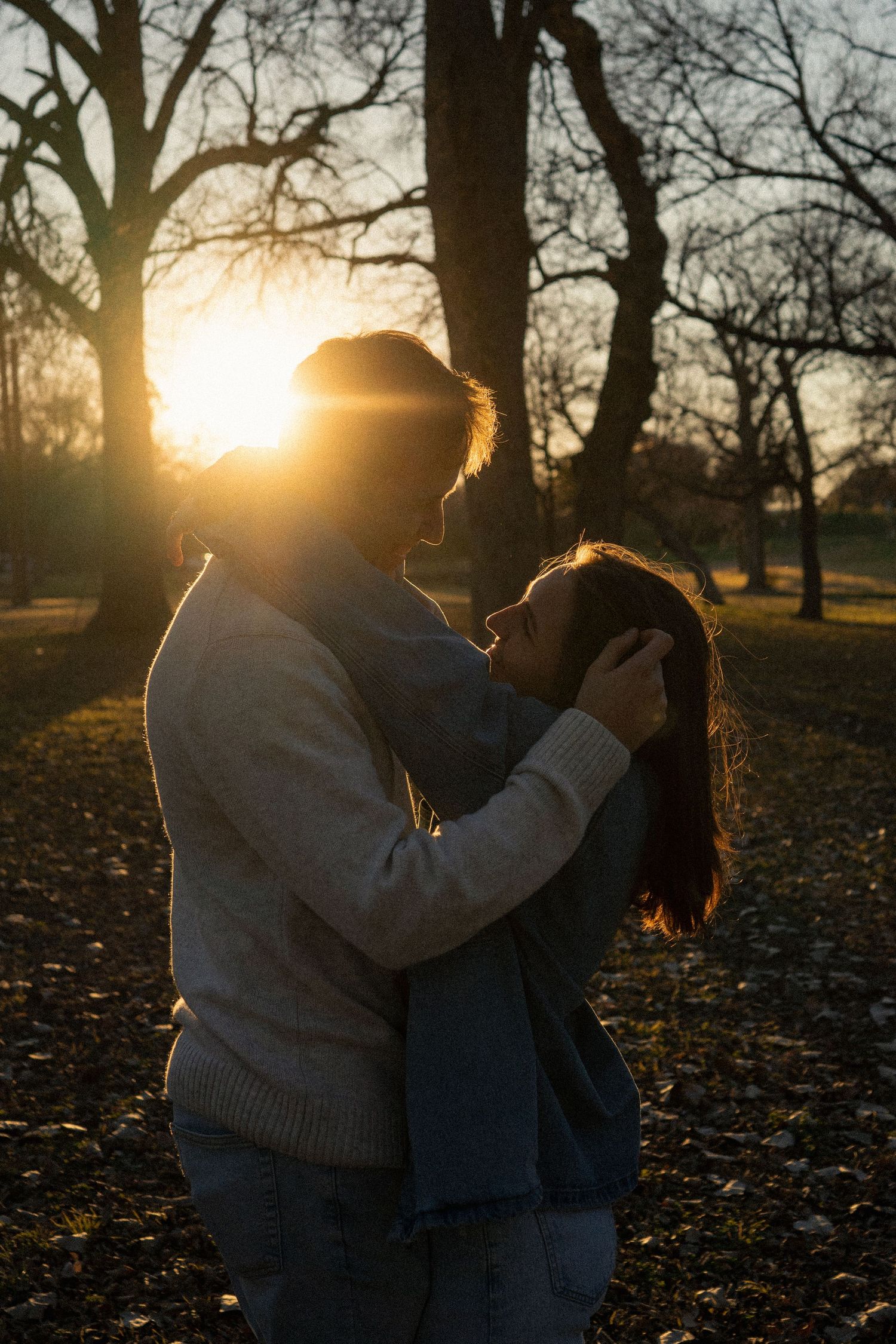 A tender embrace silhouetted against a sunset through bare trees in a park.