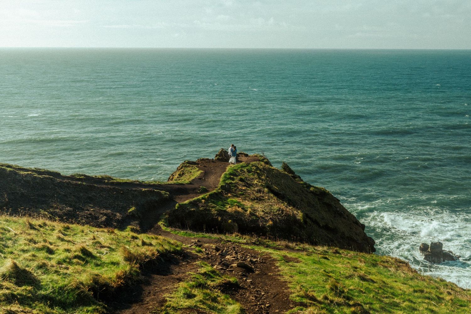 A scenic coastal path leads to a lighthouse perched on a green clifftop overlooking the turquoise ocean.