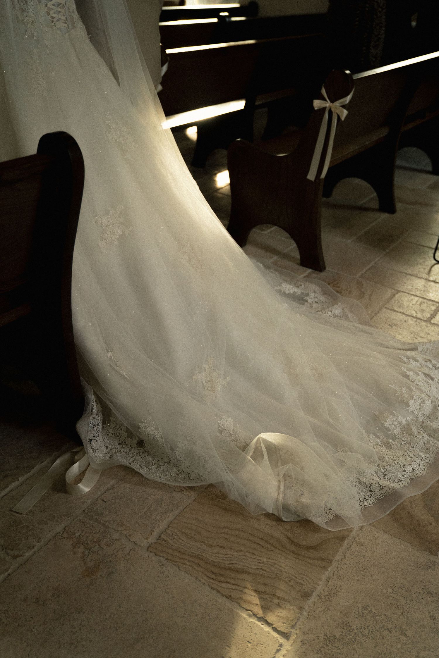 A flowing white wedding dress with delicate lace details trails along a tiled floor.