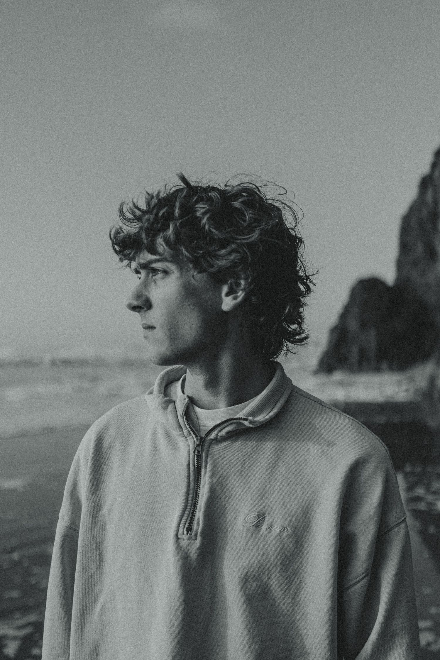 A black and white coastal portrait featuring someone in a light-colored pullover against dramatic cliffs.