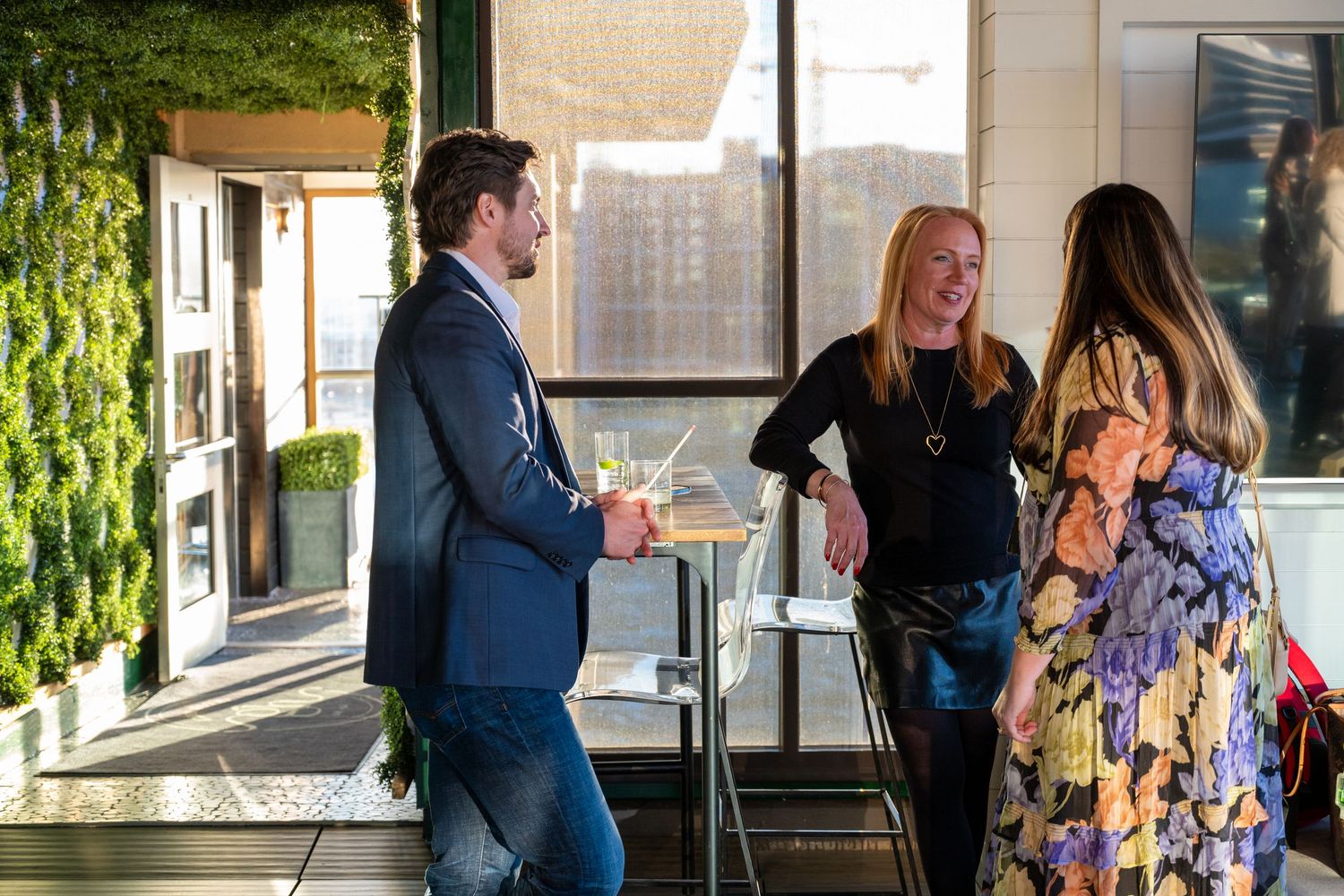 People gather and socialize at an outdoor venue with glass walls and lush green foliage in the background.