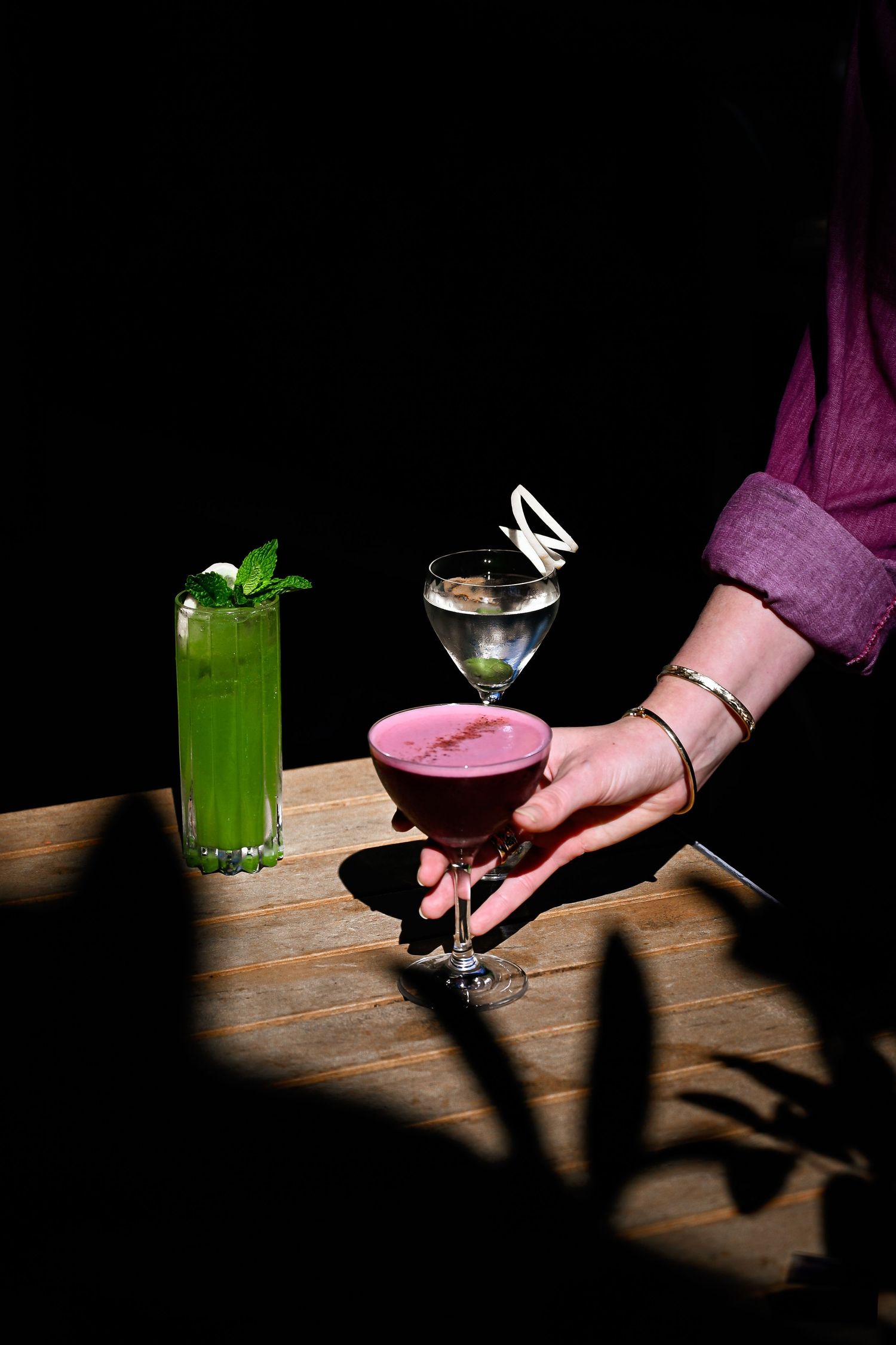 Hand holding pink cocktail glass on wooden table next to green drink, dramatic lighting casts moody shadows.