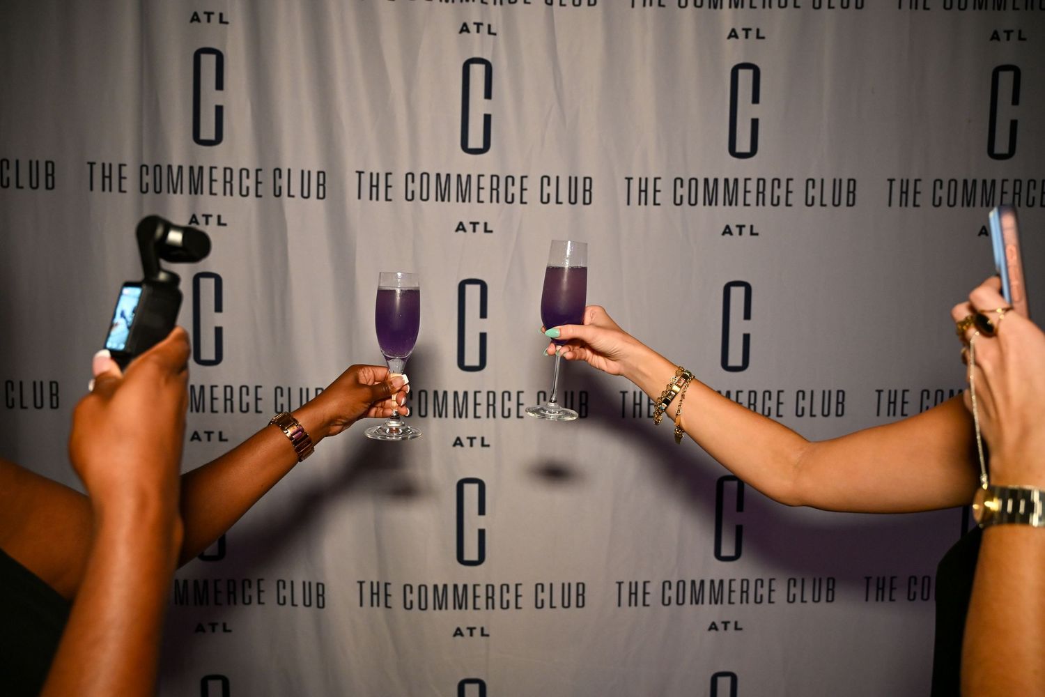 A row of hands holding up mobile phones against a branded backdrop at The Commerce Club event.