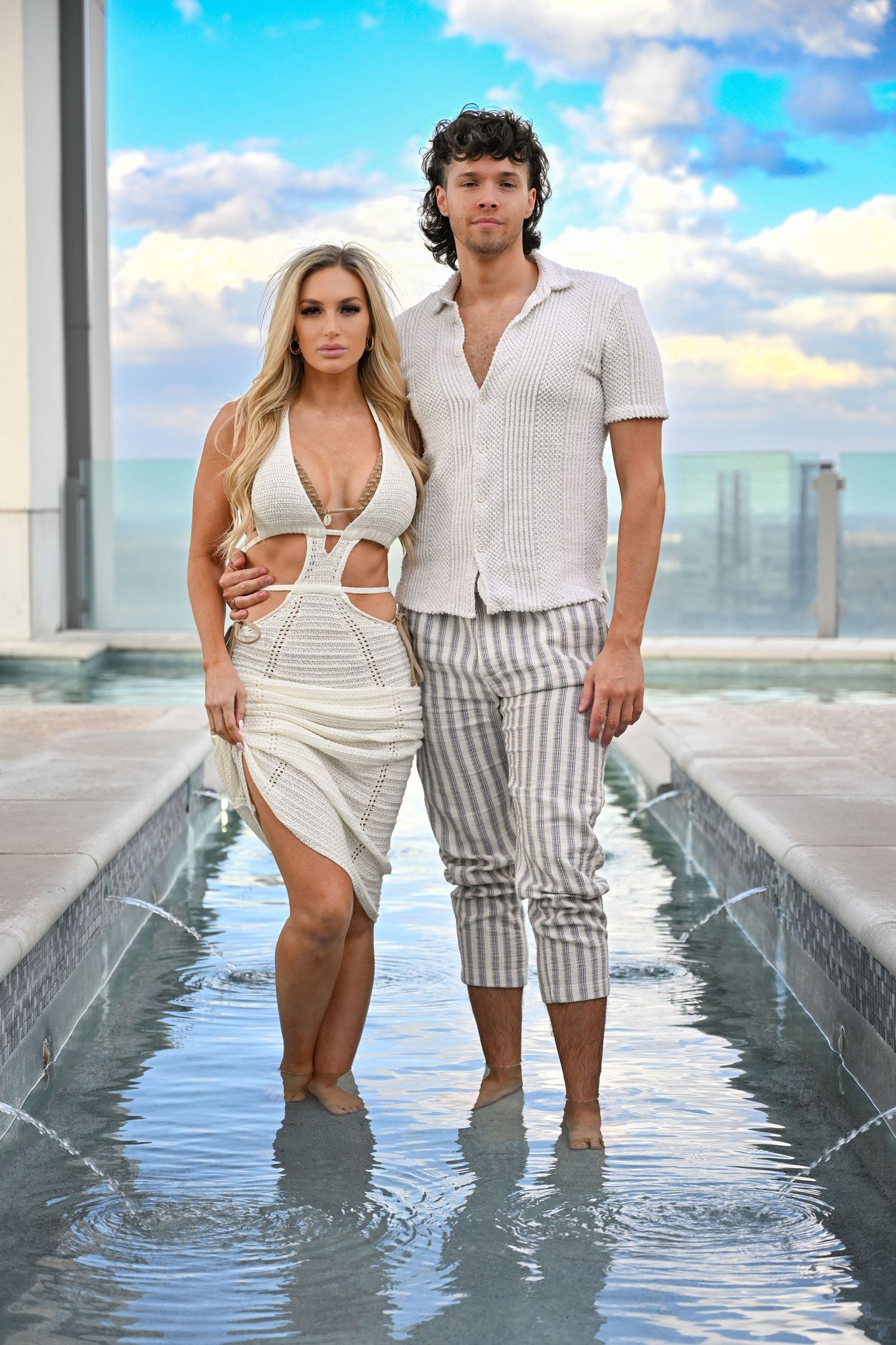 A couple in white summer outfits stands in shallow water posing together against a bright sky with clouds.