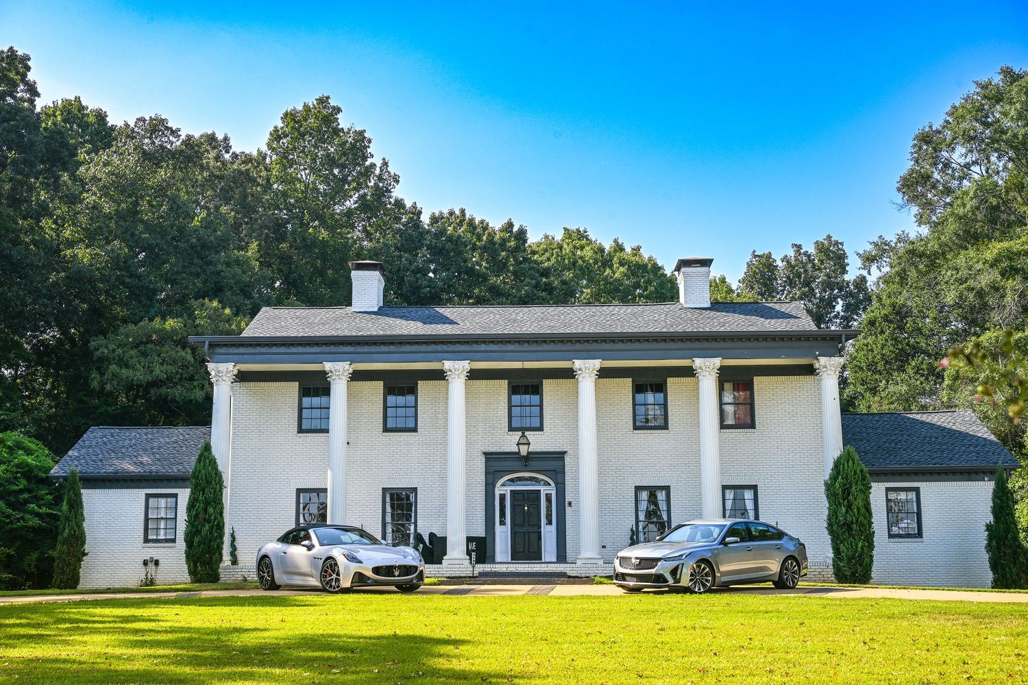 Two luxury cars parked in front of a grand white colonial mansion with tall columns and lush green lawn.