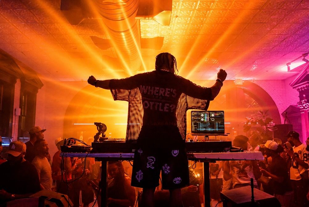 DJ performing on stage with arms outstretched, bathed in dramatic orange and yellow light beams at a packed nightclub event.