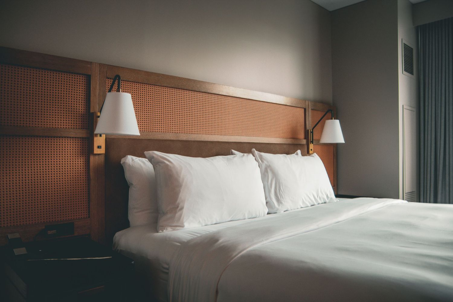 Modern hotel room interior with white bedding, wooden headboard, and wall-mounted reading lamps in warm lighting.