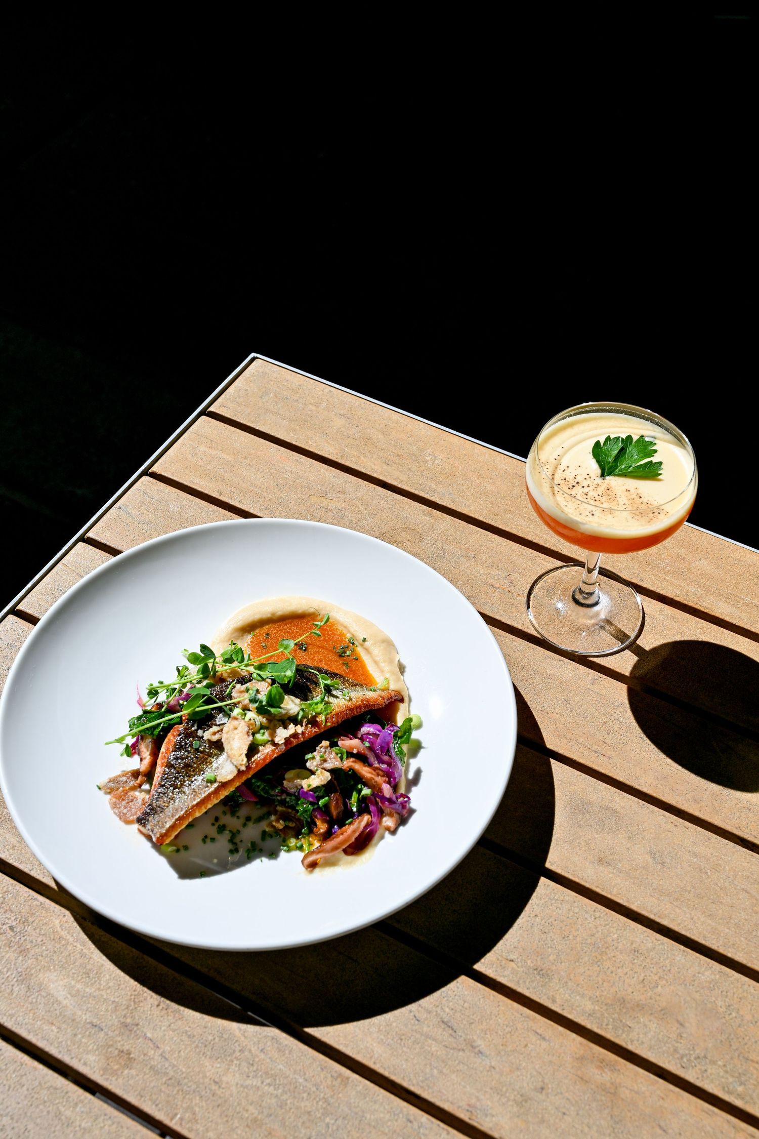 Elegant plated dish with colorful garnishes served alongside a chilled cocktail with herb garnish on wooden table.