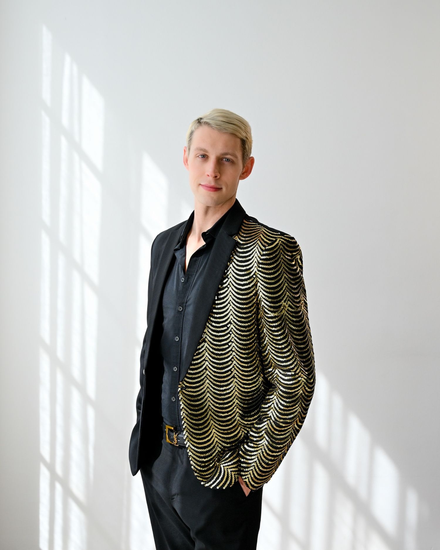 Fashion portrait of someone wearing a black shirt and striking gold patterned blazer against a white wall with shadows.