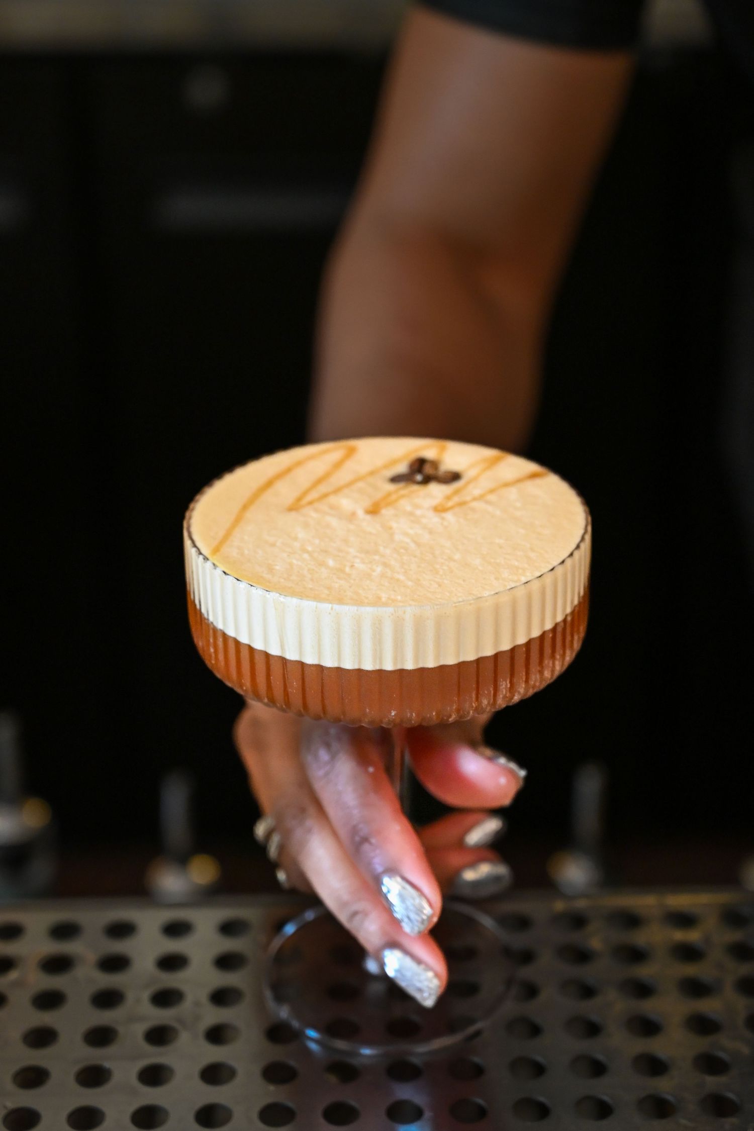 A layered espresso martini cocktail with creamy foam top is held by a hand with decorated nails on a bar mat.