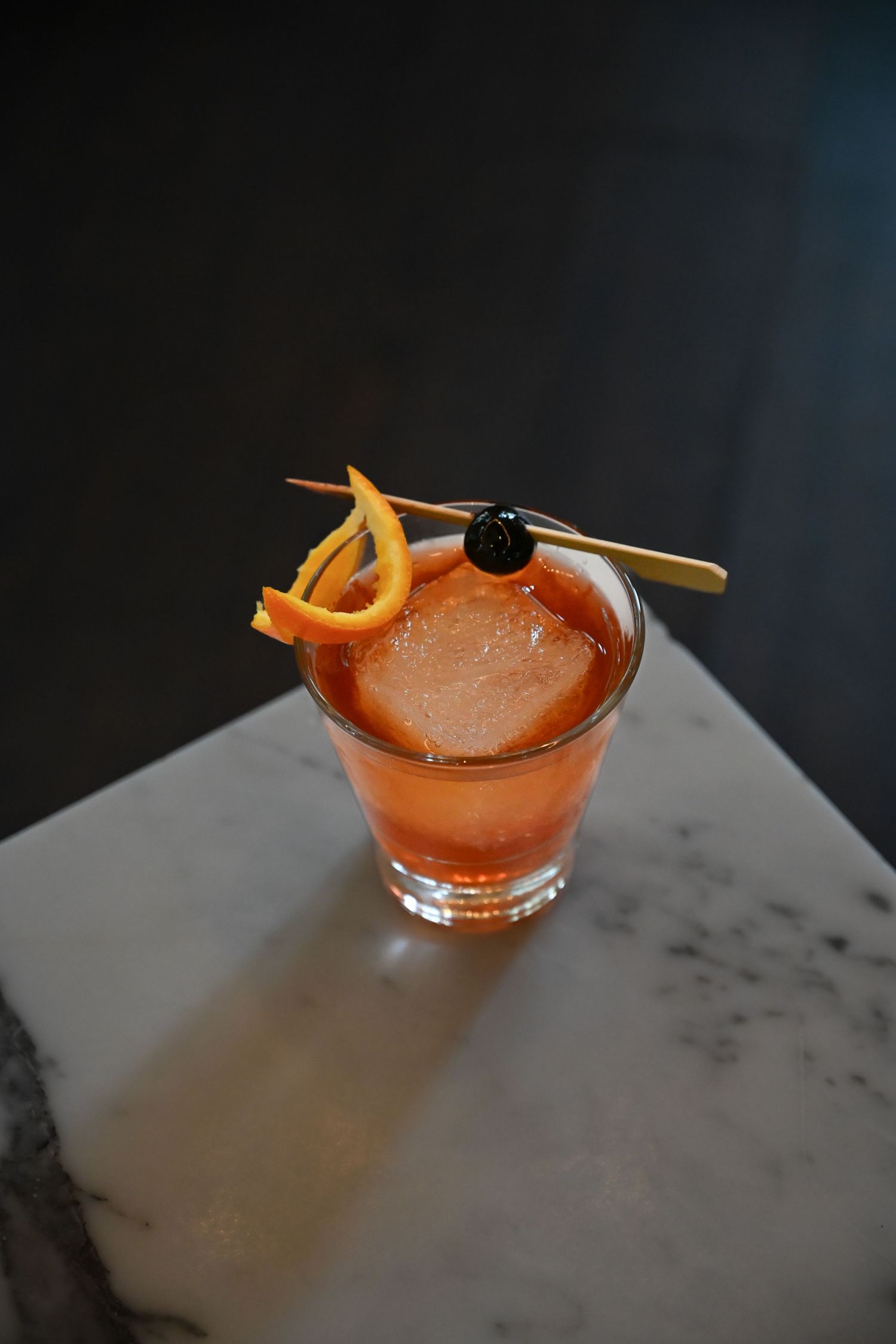 A craft cocktail garnished with an orange peel and dark cherry sits on a marble bar counter.