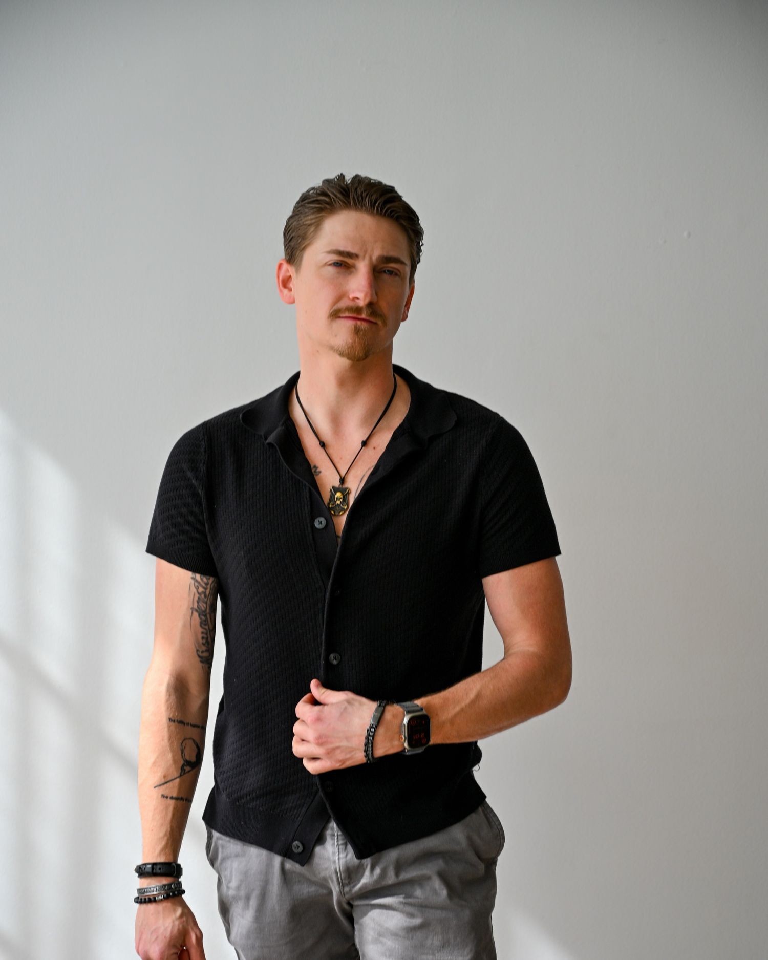 Man in black button-up shirt with tattoos and necklace posing confidently in a bright studio setting.