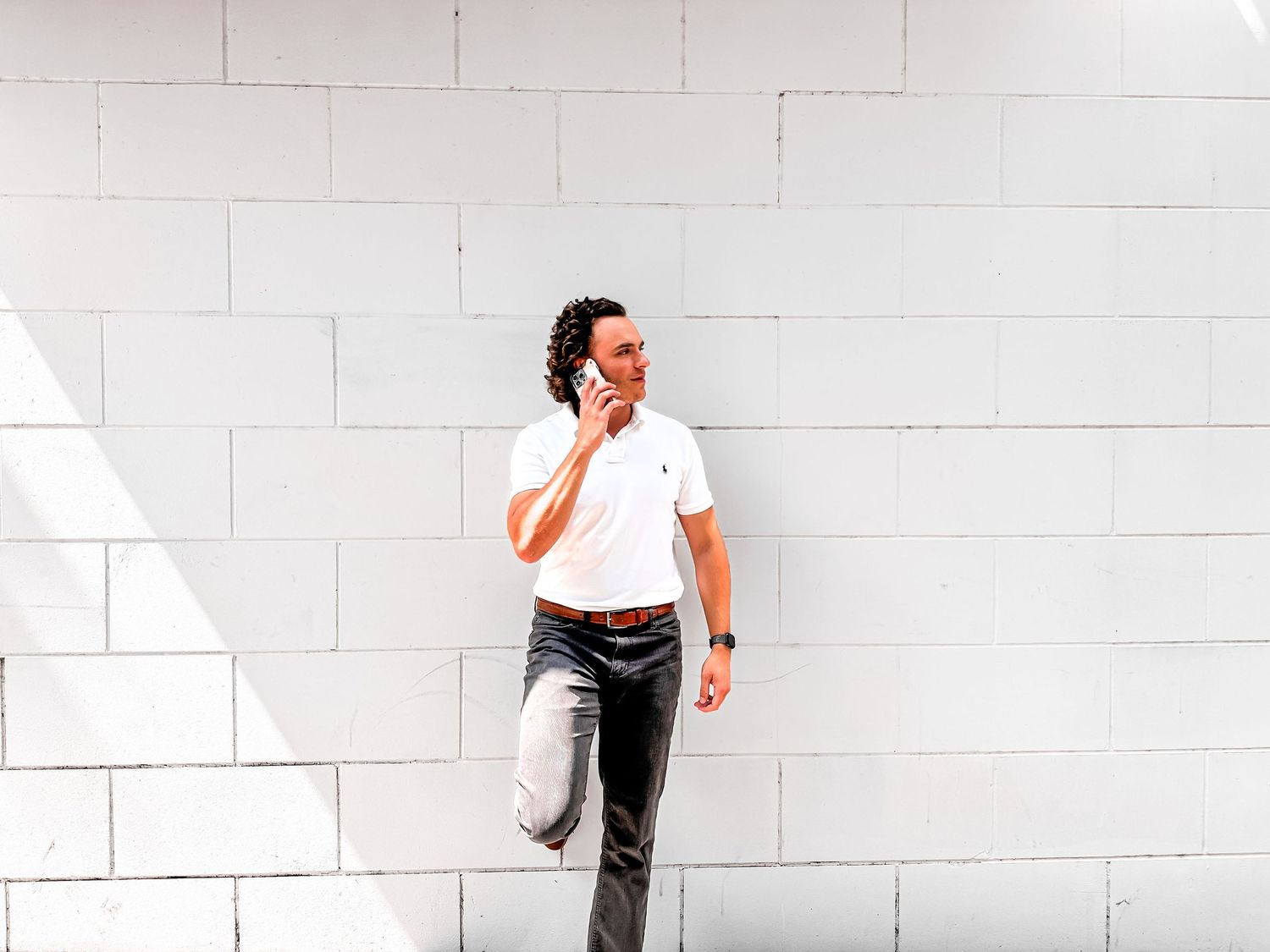 Person in casual attire leaning against white brick wall while talking on phone.