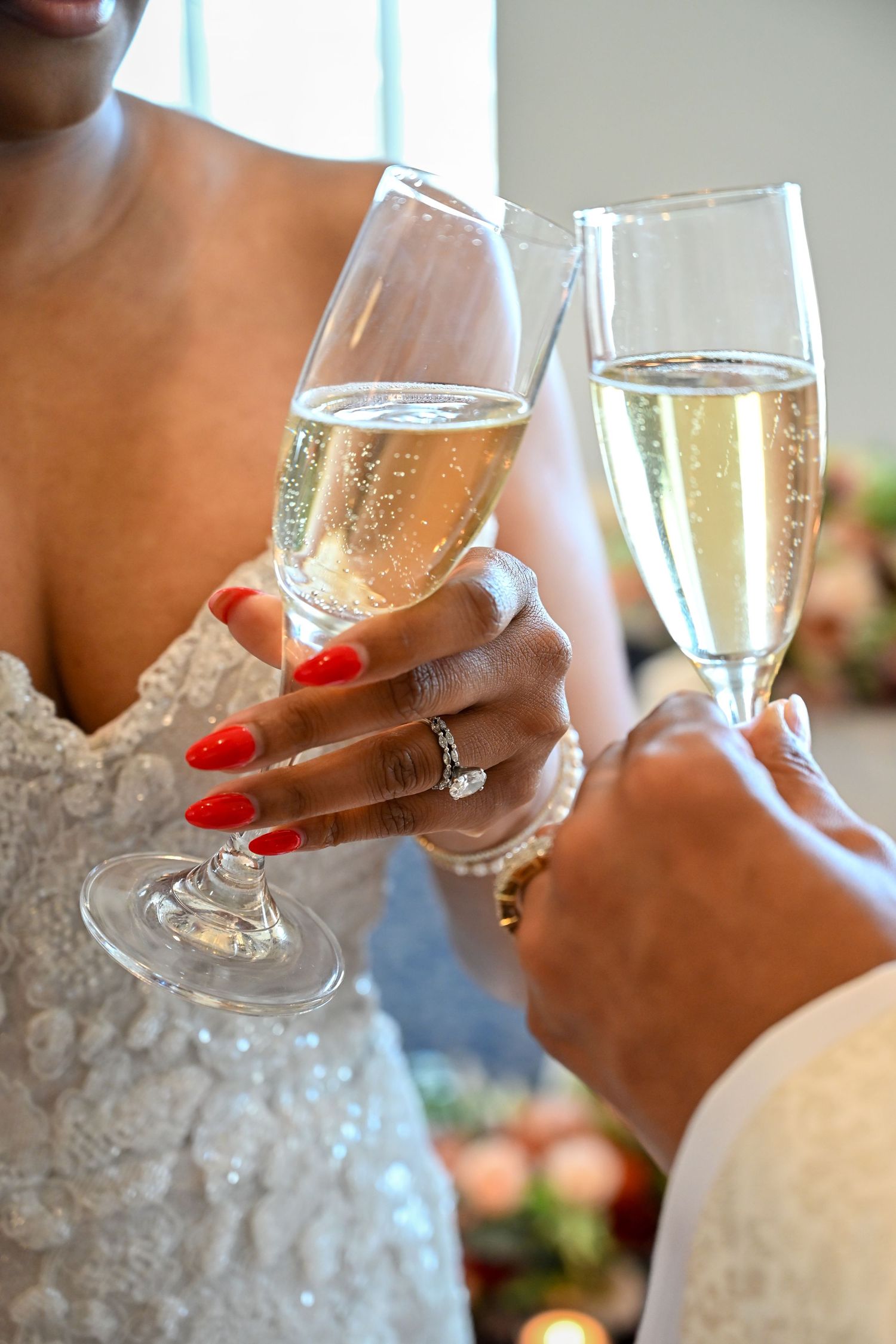Two champagne flutes clink together in a toast, with red manicured nails and a white lace dress visible in the frame.