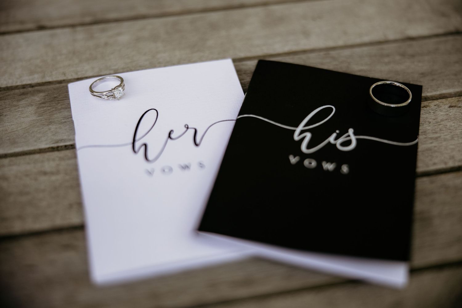 Black and white wedding vow books labeled 'his' and 'hers' arranged on wooden surface.