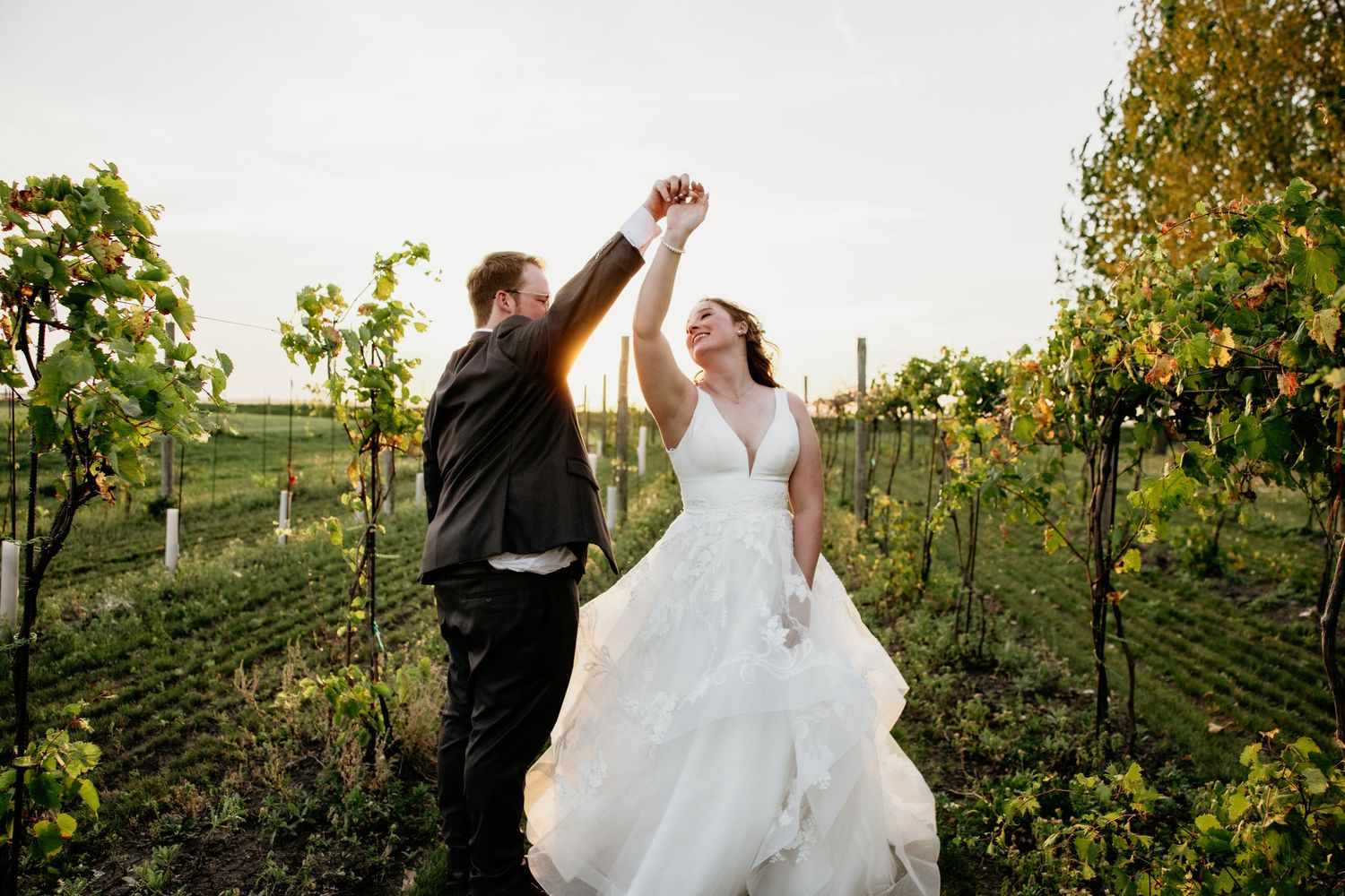 A couple dances joyfully among vineyard rows at sunset during their wedding celebration.